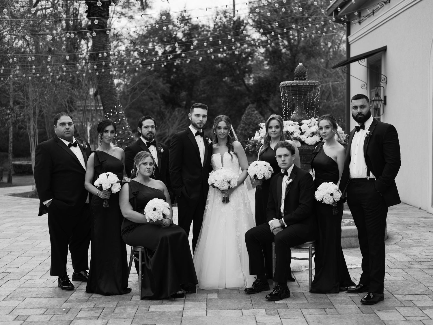 Wedding party in formal attire poses together outdoors on a sunny day.