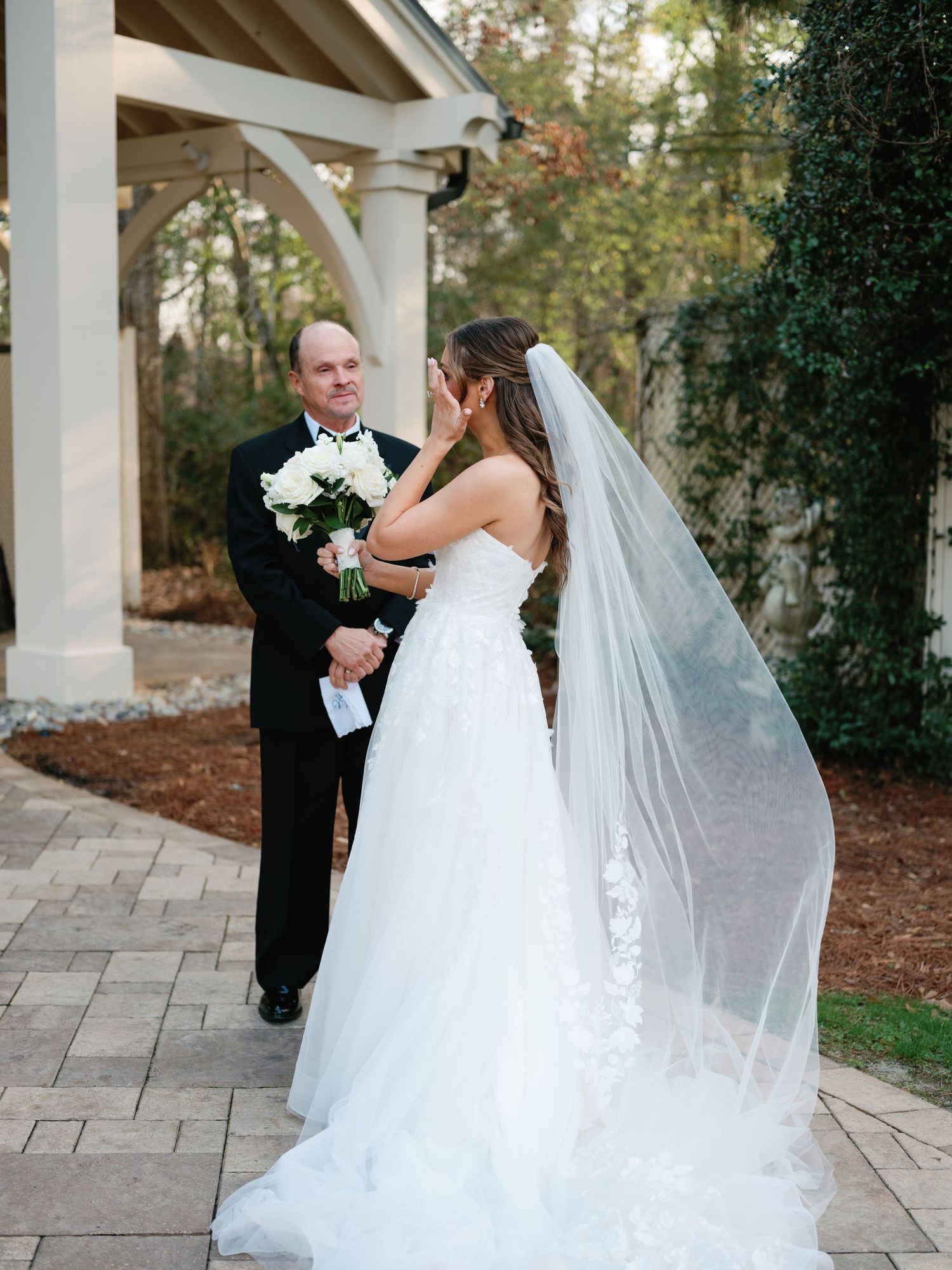 A wedding ceremony moment under a white pergola with flowing veil and white bouquet.