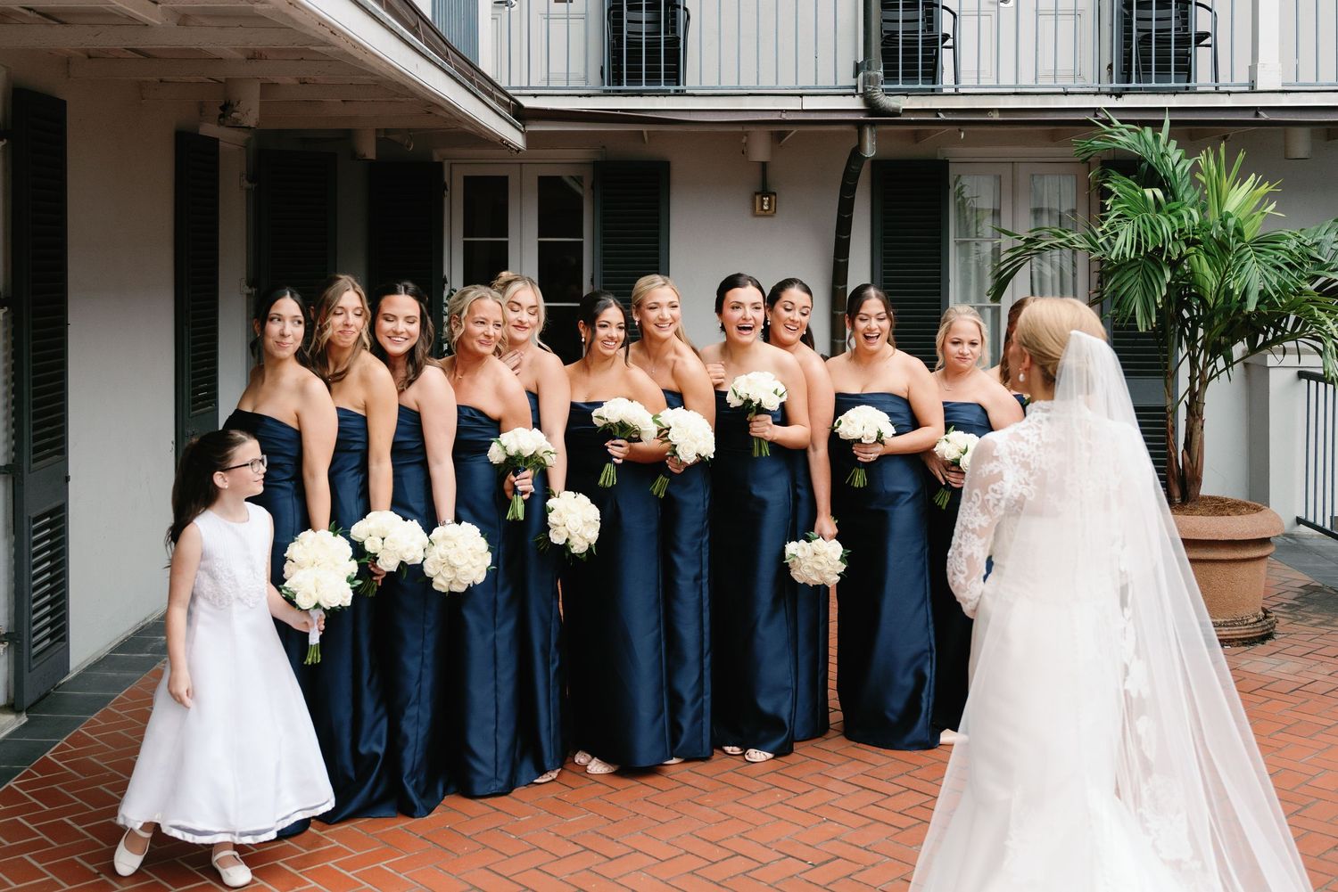 Wedding party in navy blue dresses lined up along a brick courtyard holding white bouquets.