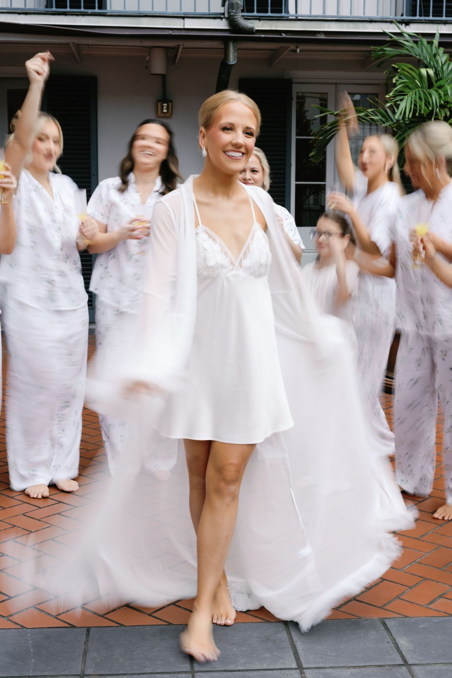 Bridesmaids in white dresses dancing and celebrating together on a tiled patio.