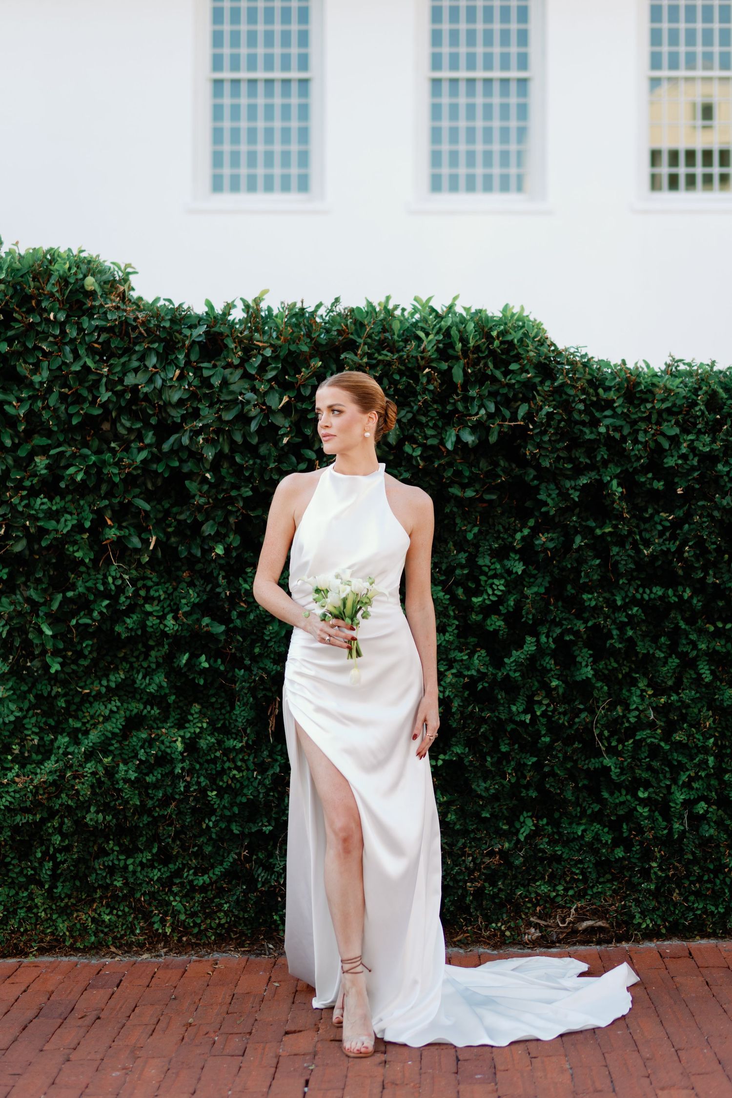 Elegant bride in flowing white gown poses on brick pathway by hedge wall.