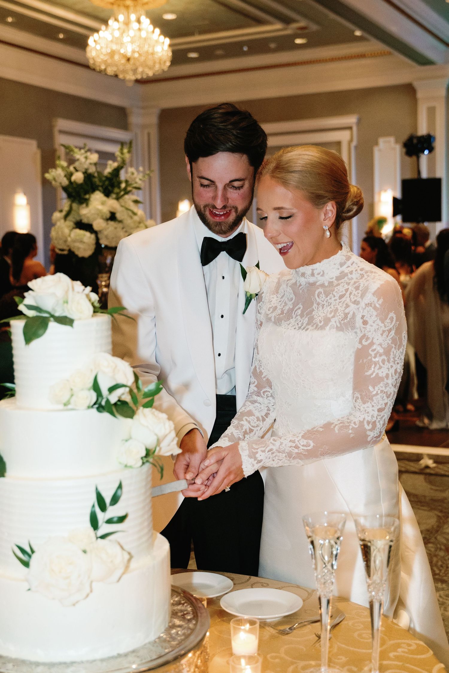 A couple in formal wedding attire cuts into their elegant white tiered cake adorned with greenery at an elegant reception.