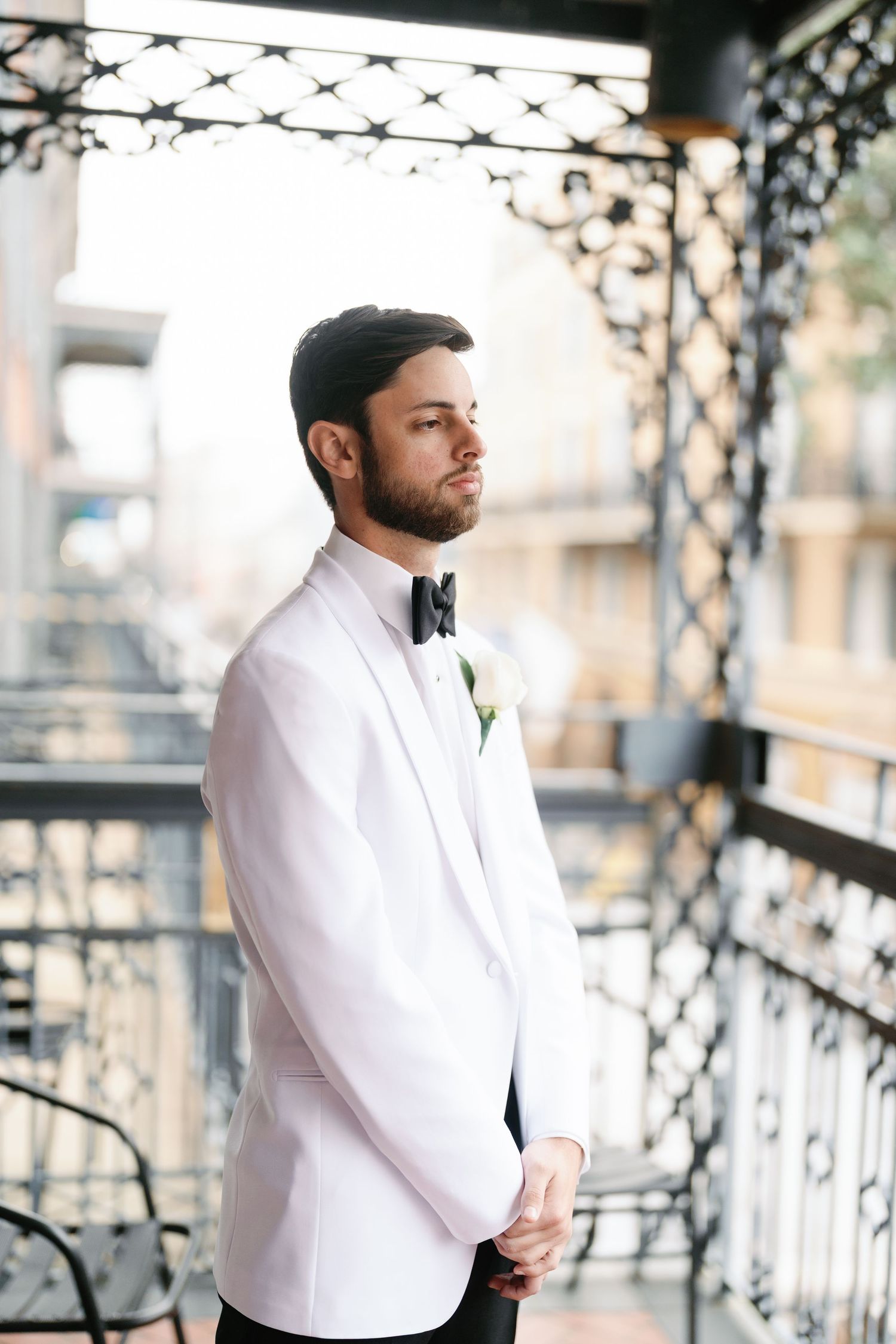 Groom in white dinner jacket and black bow tie stands on ornate balcony.