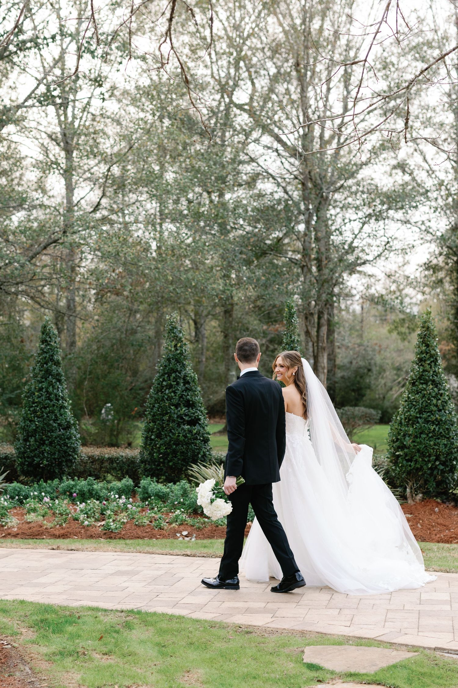 A couple in wedding attire walks along a stone path surrounded by manicured topiary trees and landscaping.