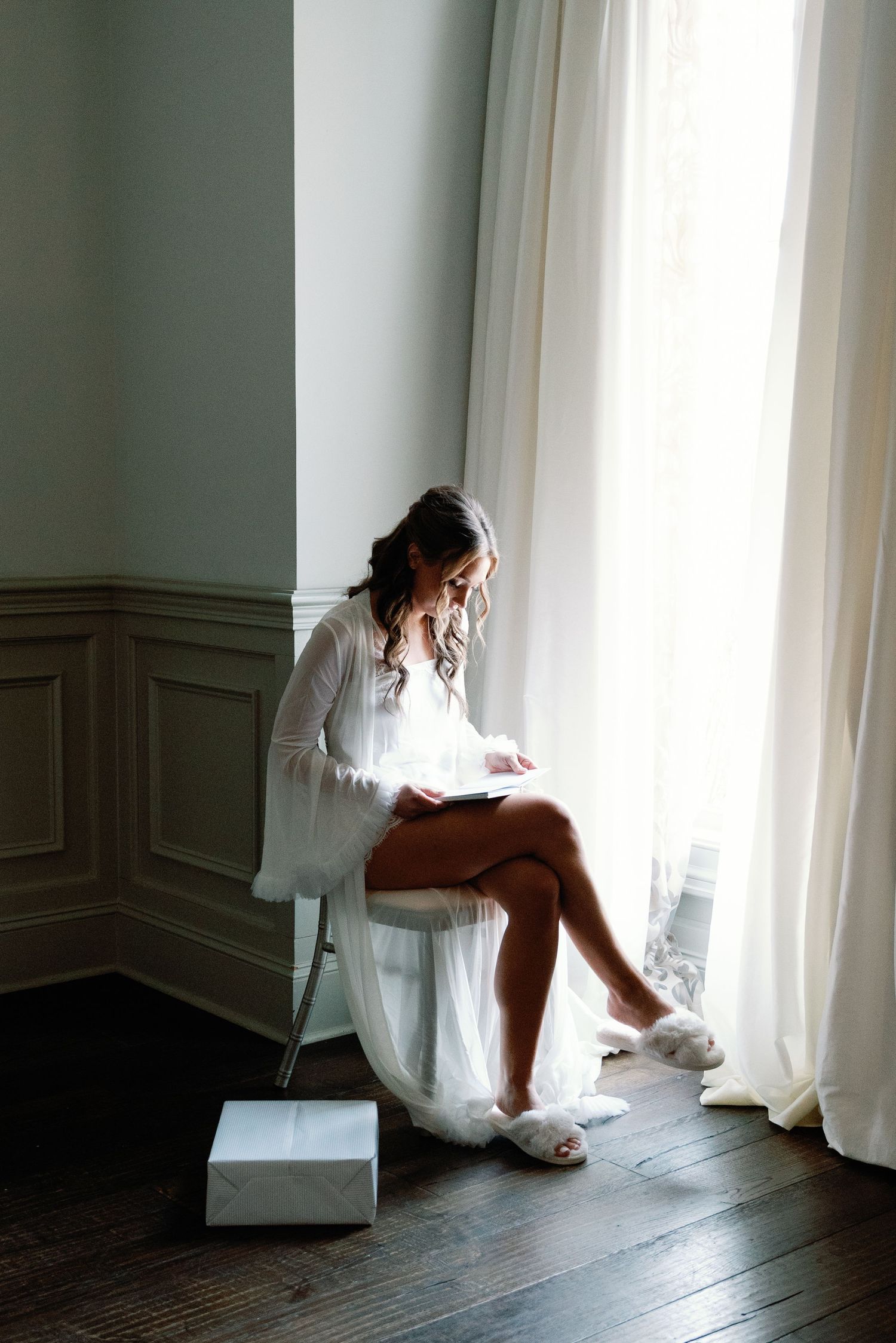 Soft morning light streams through white curtains in an elegant room with wainscoting and hardwood floors.