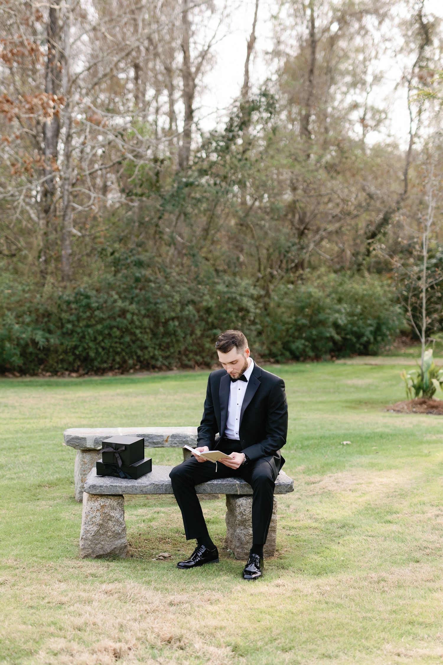 A person in formal attire sits alone on a rustic stone bench in a garden setting.