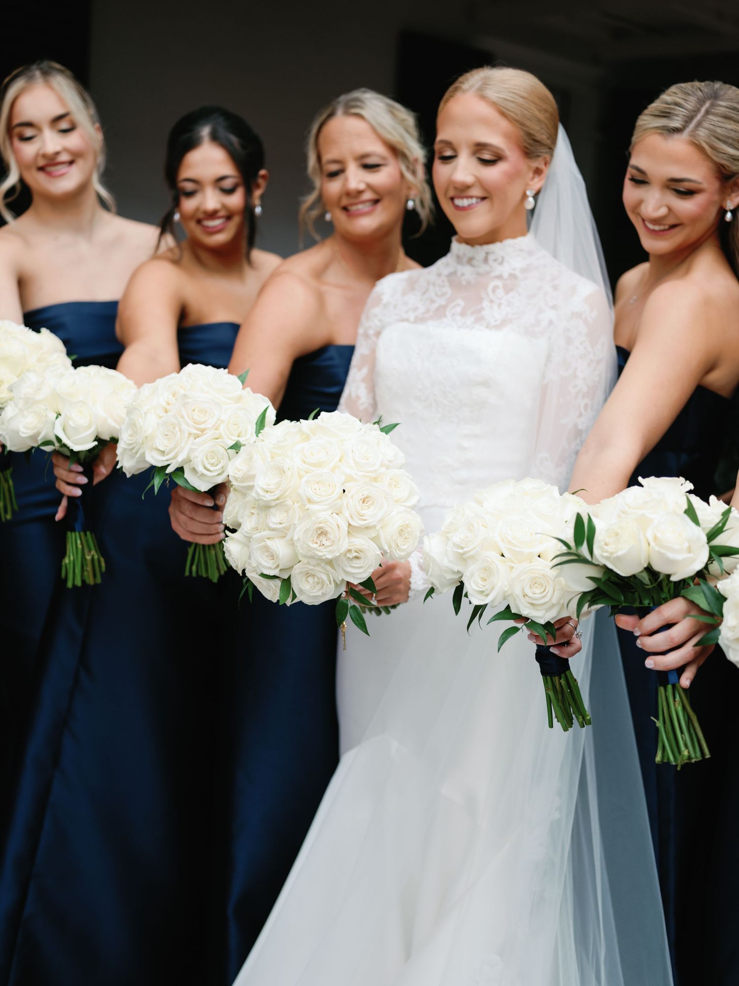 Bridesmaids in navy dresses hold white rose bouquets while standing with the bride.