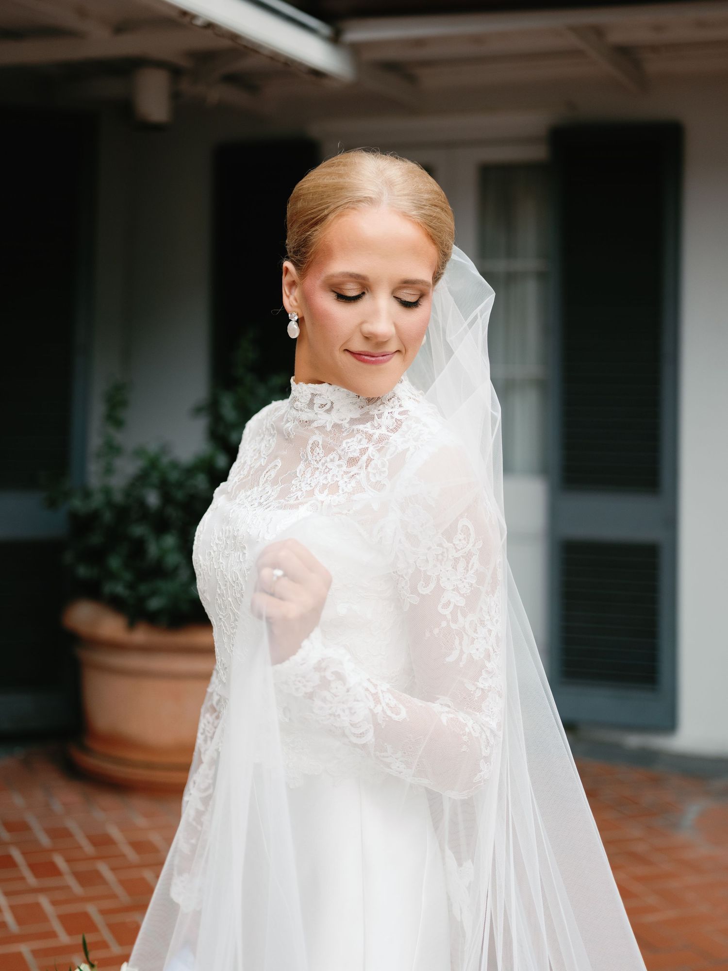 Bride in high-neck lace wedding dress poses gracefully with flowing veil against white exterior.