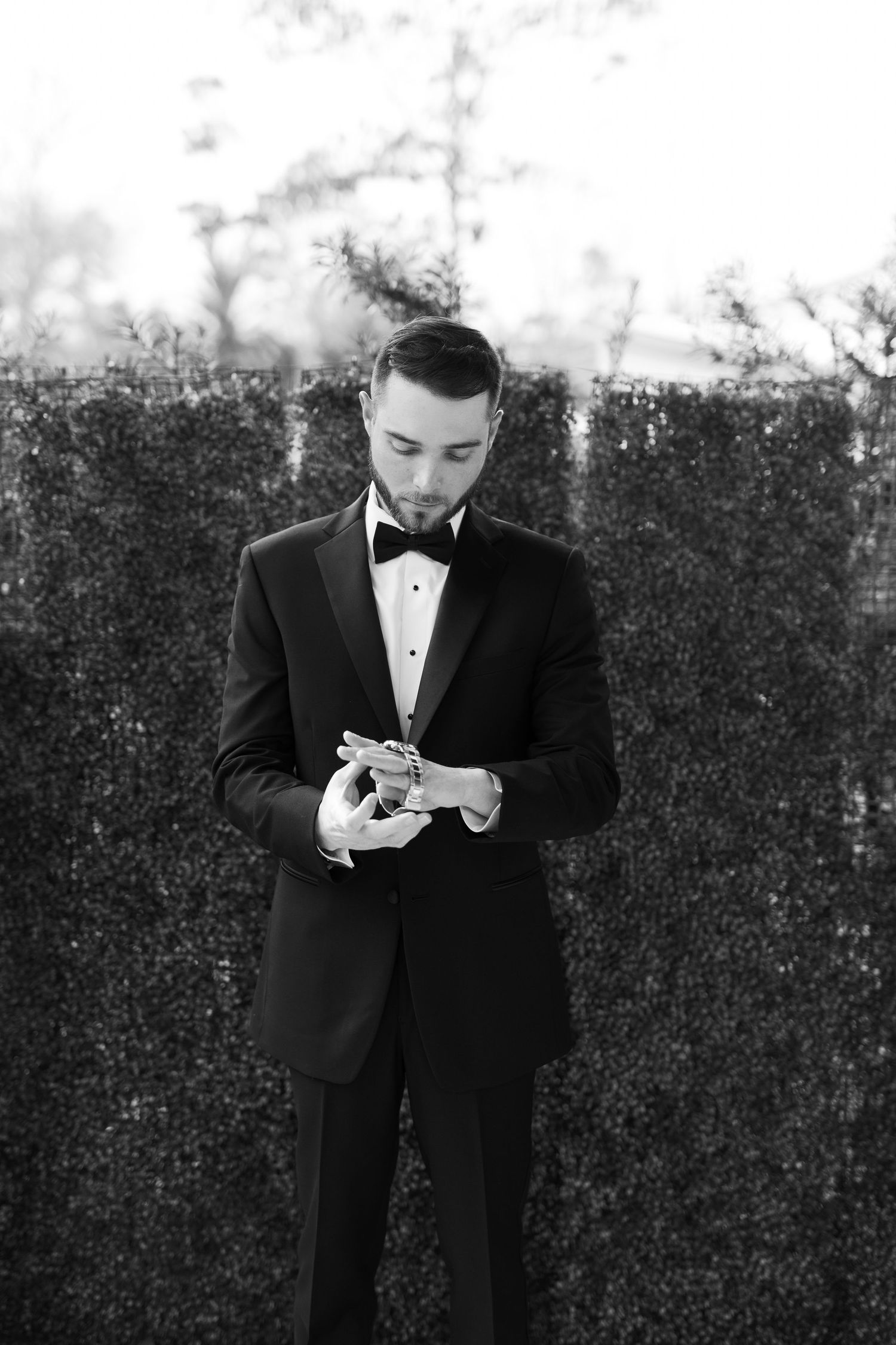 A person in a black tuxedo and bow tie looks down at their mobile phone against a dark background.