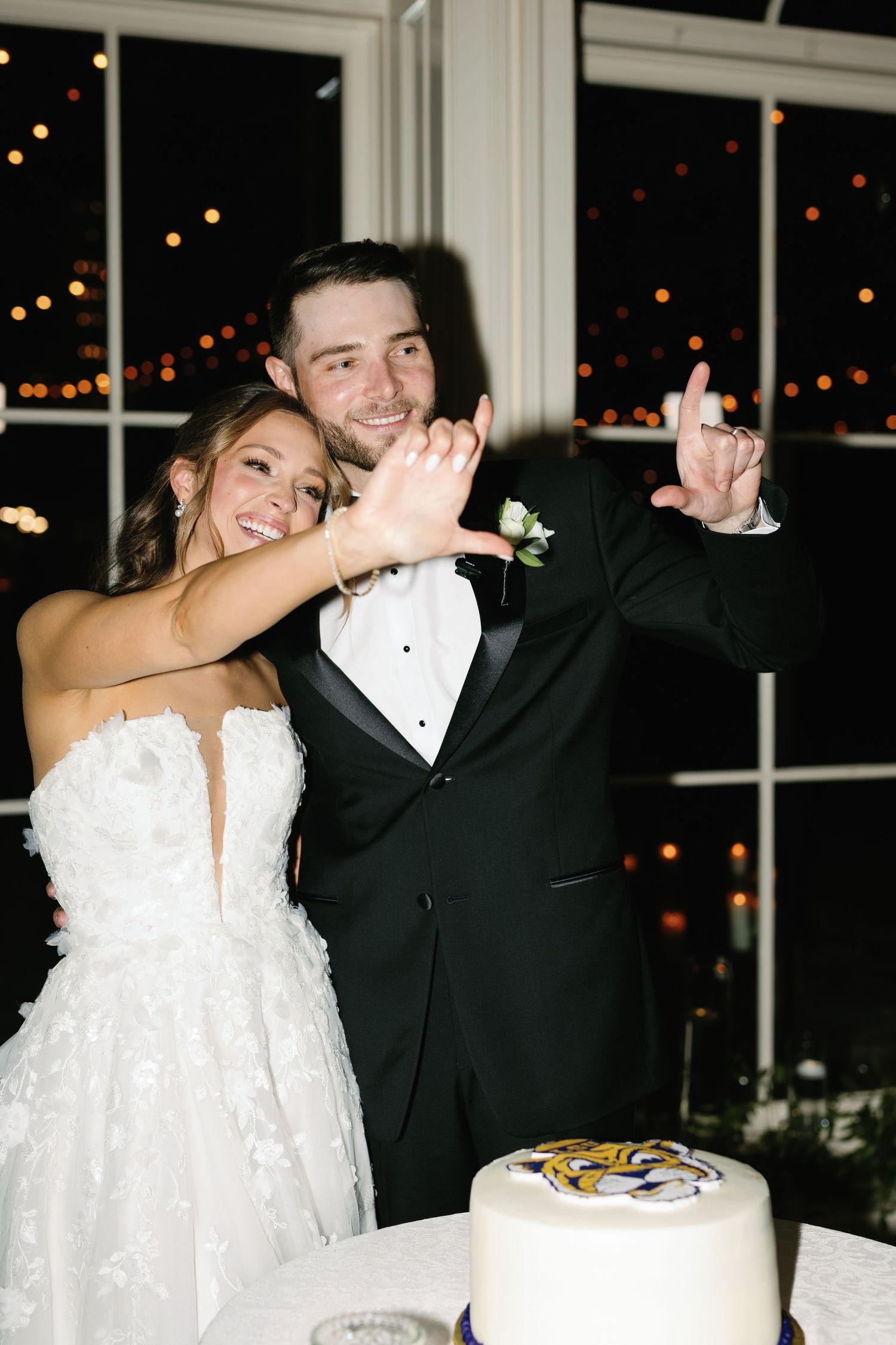 Newlyweds playfully cut their wedding cake together at an evening reception with twinkling lights.