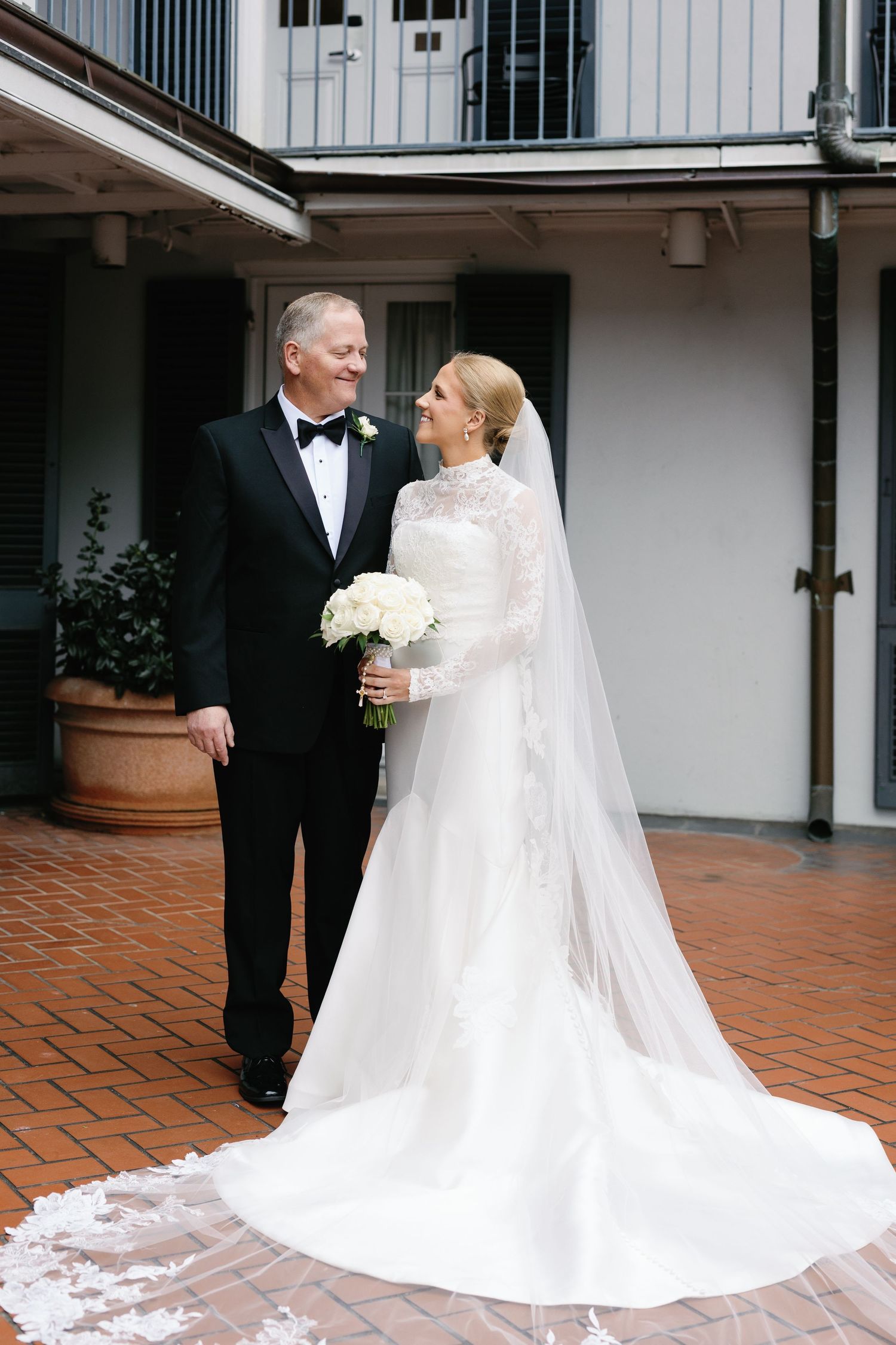 Elegant bride in a lace long-sleeved gown with flowing train poses with companion on red brick patio.
