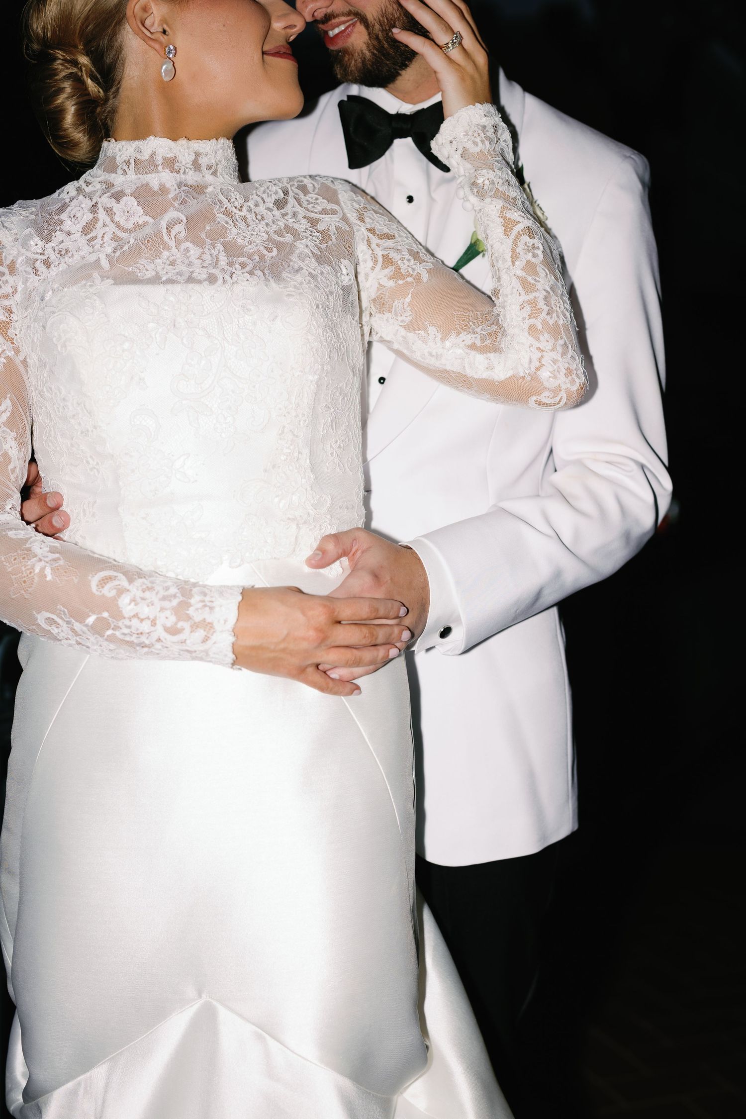 Close-up of bride in lace gown embracing groom in white tuxedo jacket.