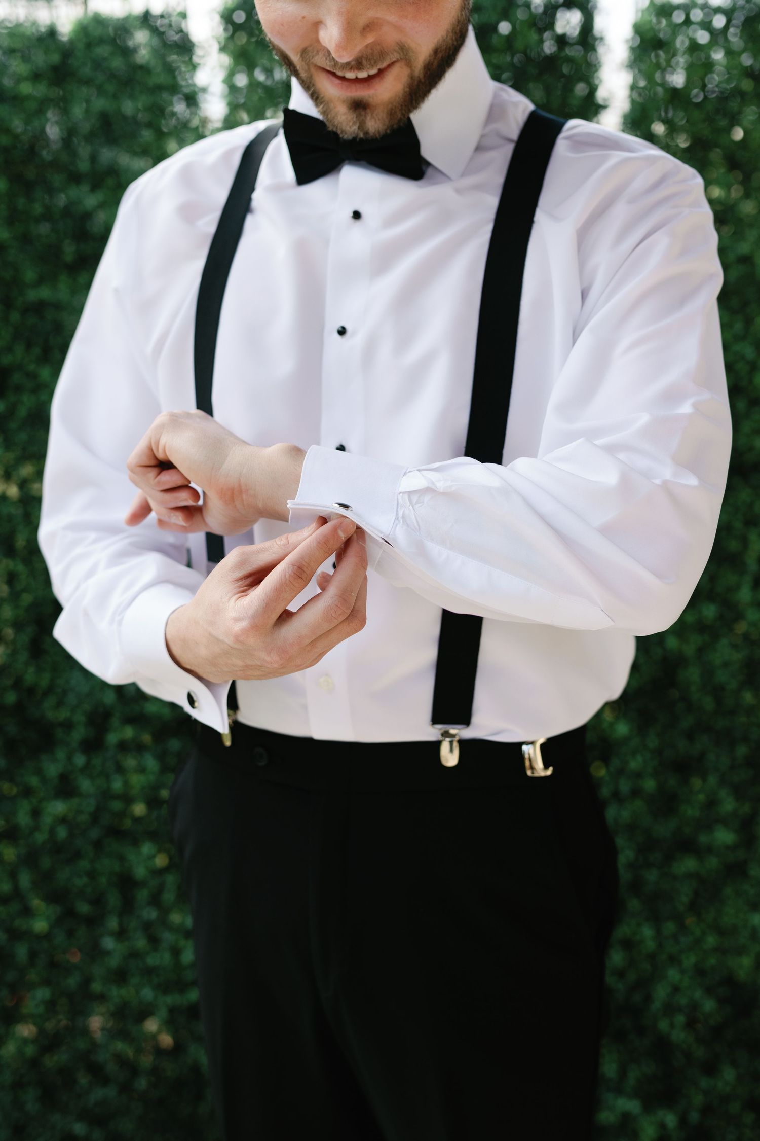 A person in formal attire adjusts their cufflinks while wearing a white dress shirt and black suspenders.