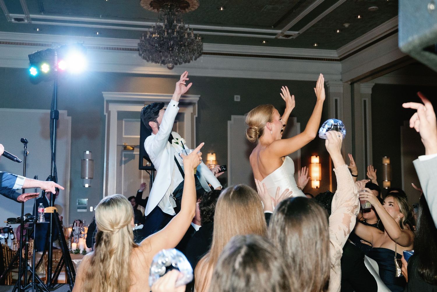Wedding reception guests raise their hands in celebration on a dimly lit dance floor.