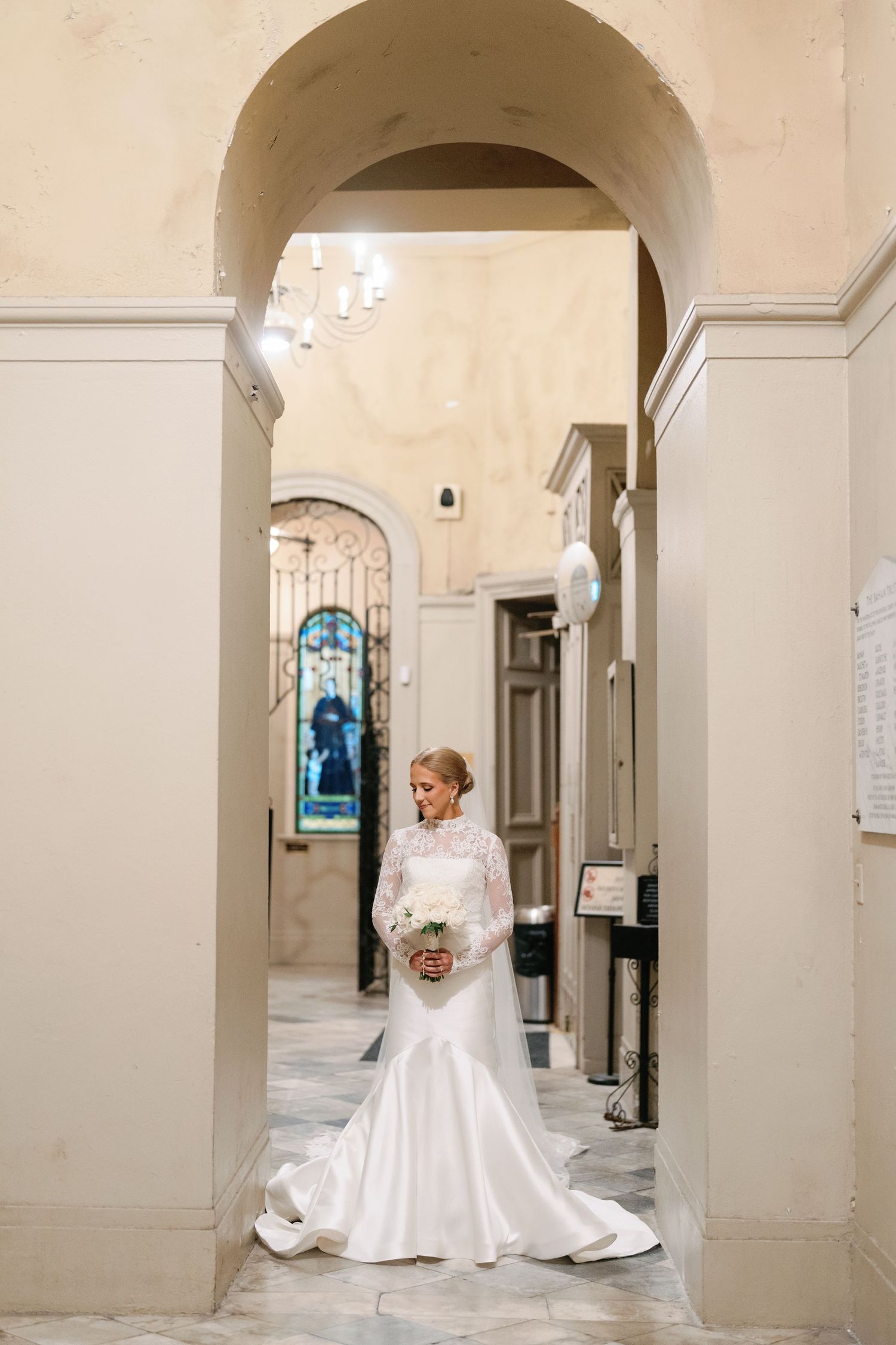 Elegant bride in long-sleeved white wedding gown poses under arched hallway.