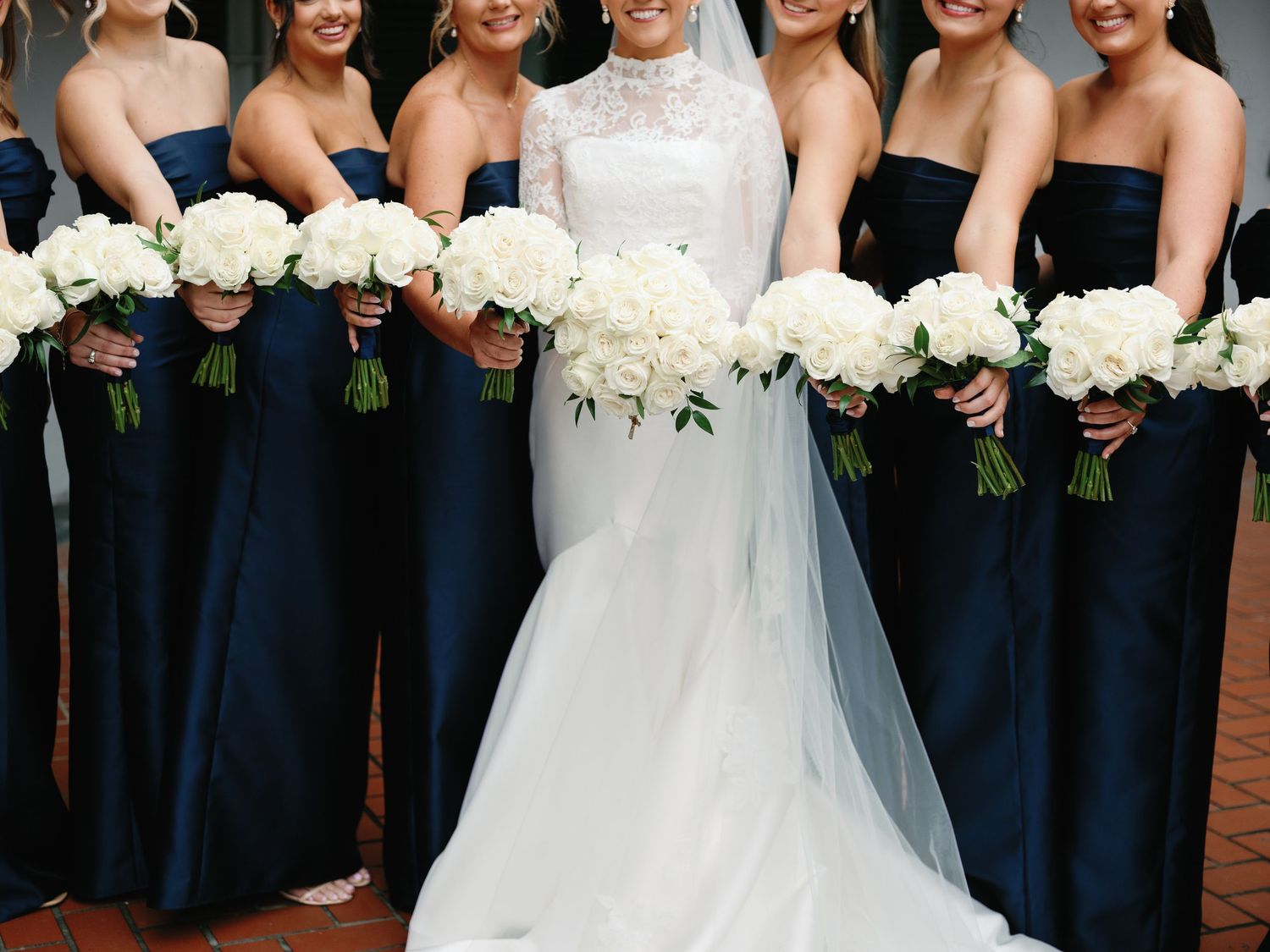Wedding party members in navy blue bridesmaid dresses stand together holding white floral bouquets.