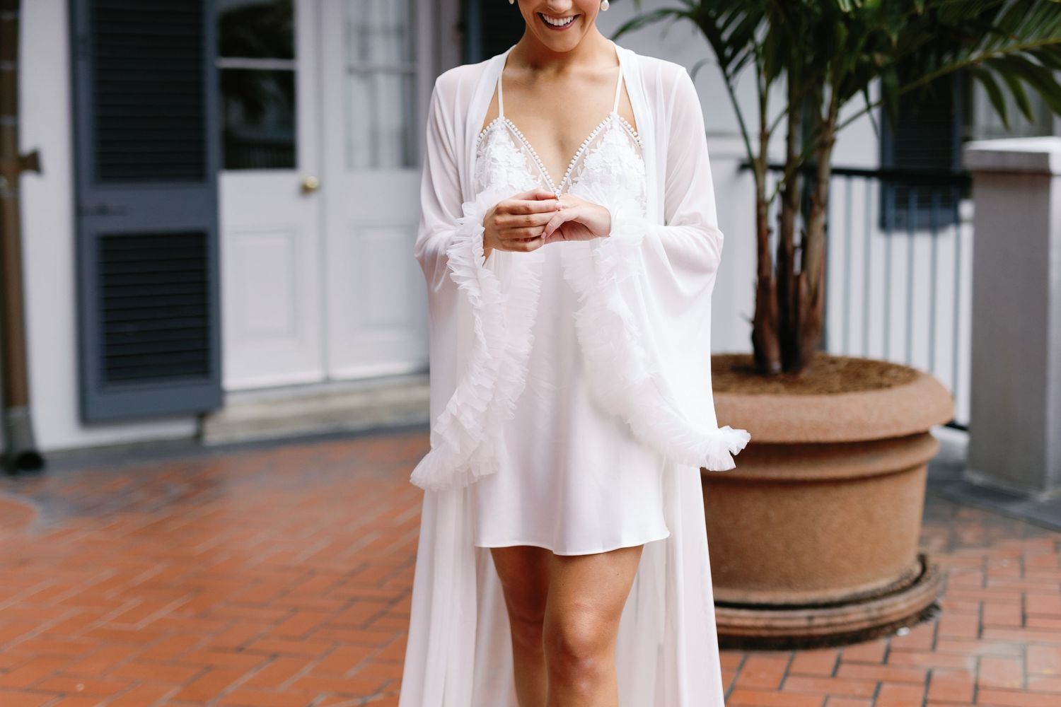 A person in a white flowing robe and short dress holds a drink while standing on terracotta tiles near a potted palm.