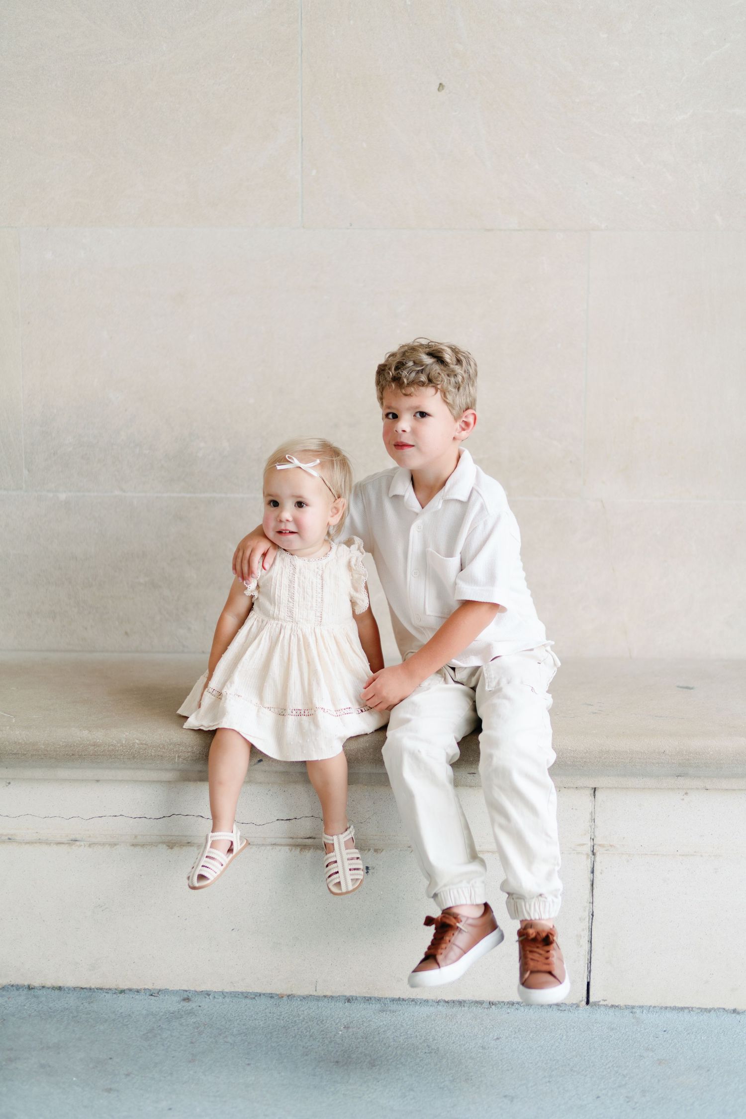 Two children in white outfits sit together on a ledge against a light colored wall.