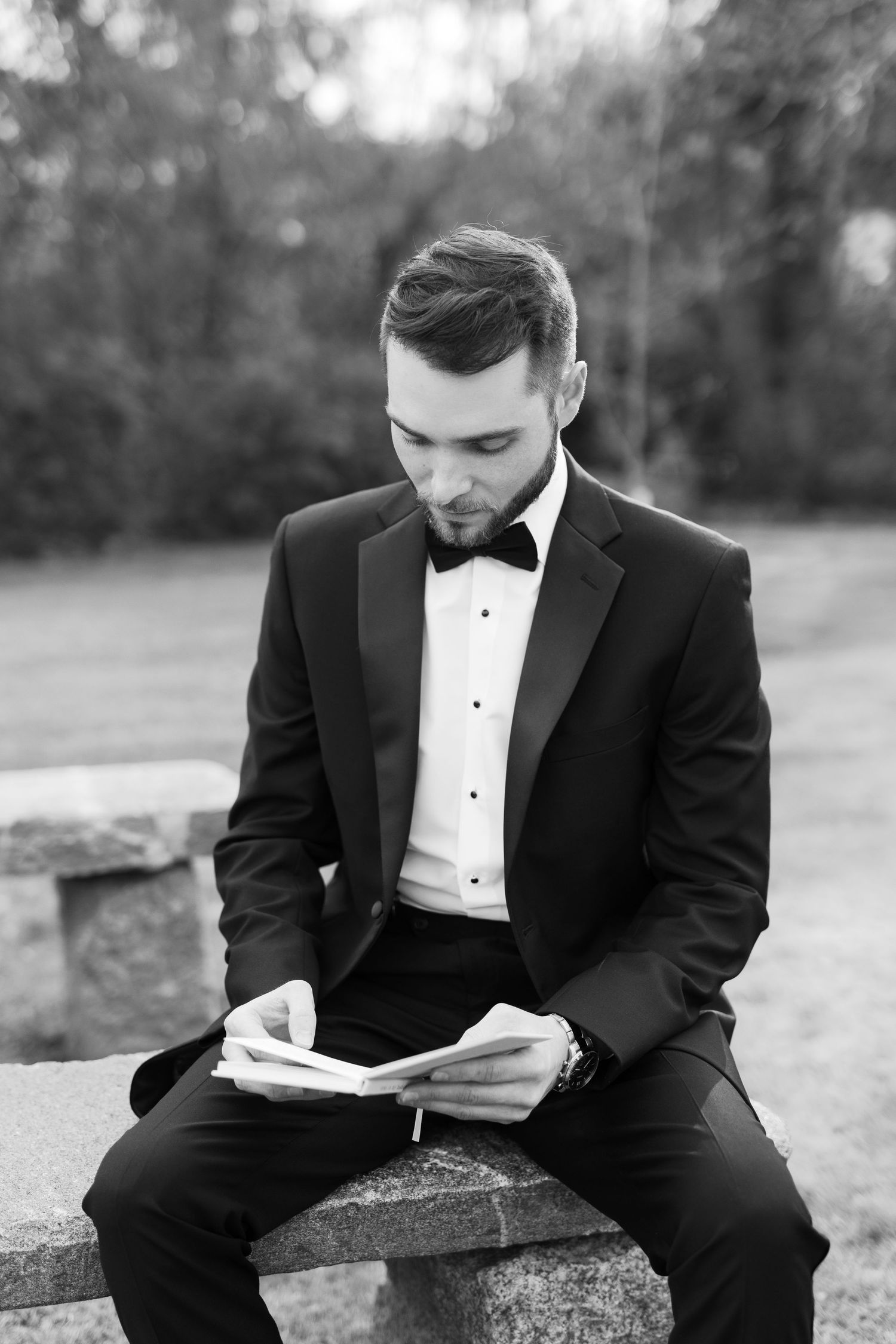 A groom in formal attire reads while seated outdoors in black and white photography.