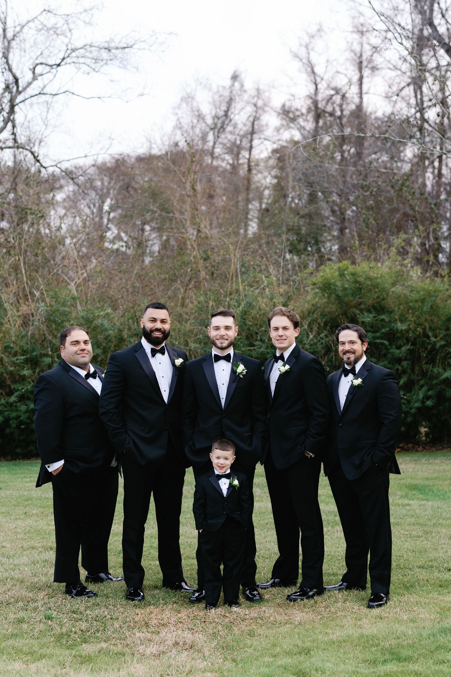 A group of groomsmen in black tuxedos pose together on a lawn for formal wedding photos.