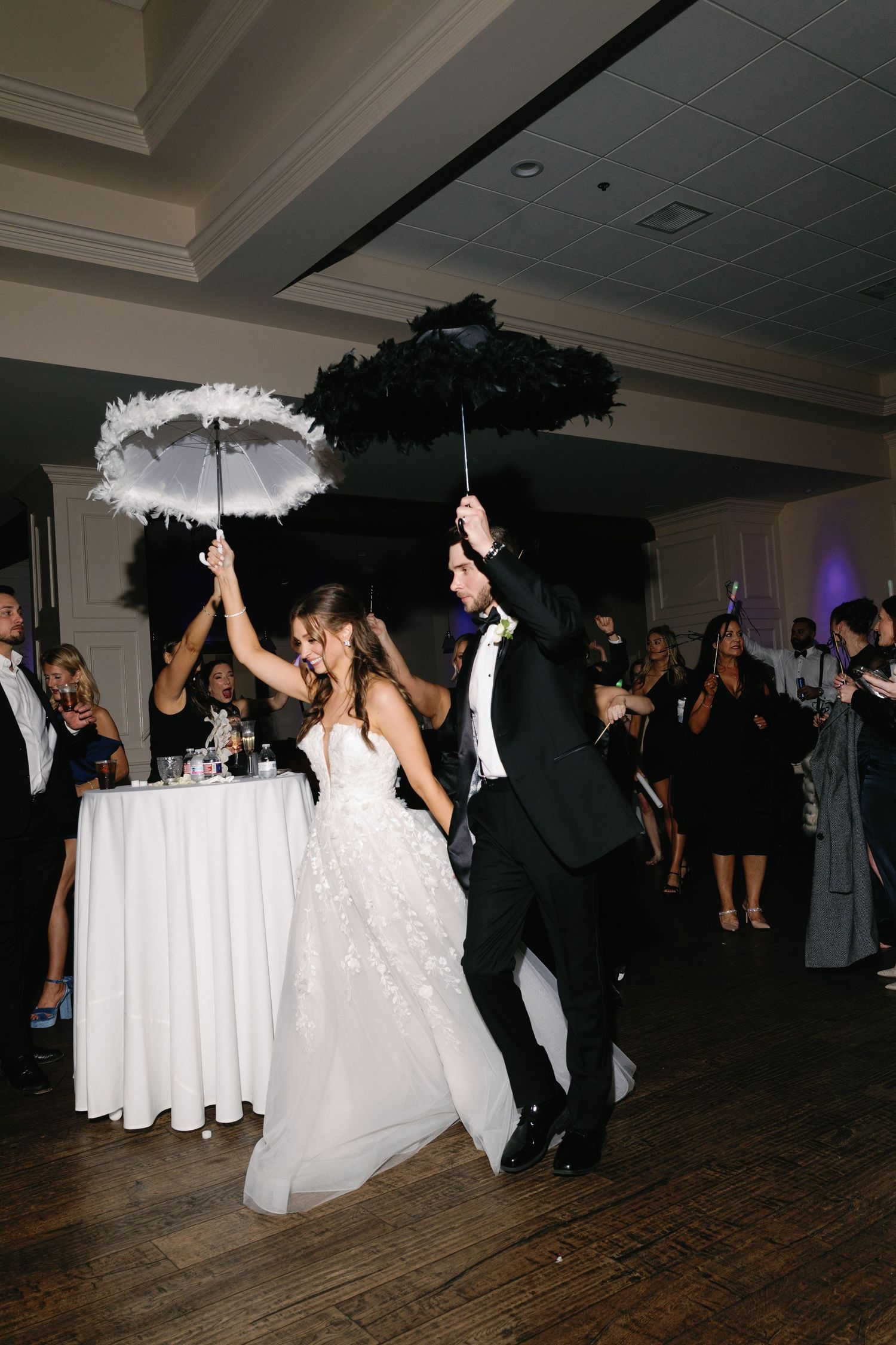 Wedding reception guests dance and celebrate on a dark dance floor with white tablecloths visible in the background.