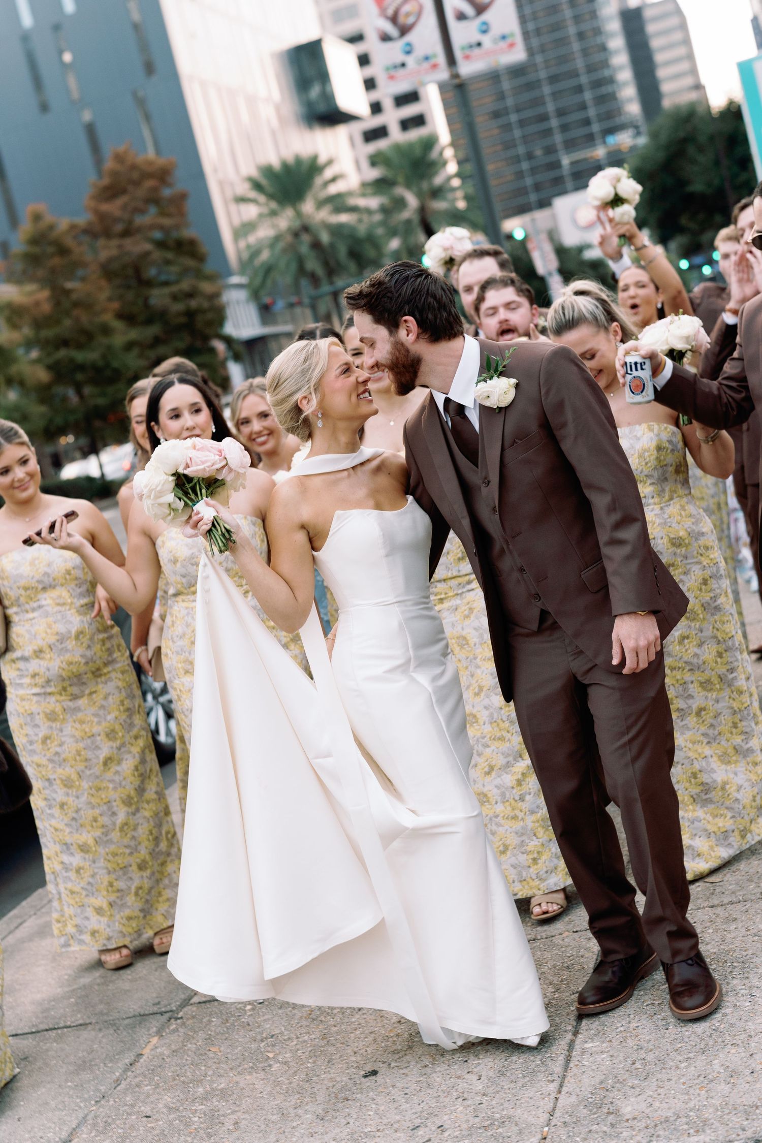 Wedding party celebrates outdoors with bridesmaids in floral dresses and groomsmen in brown suits.