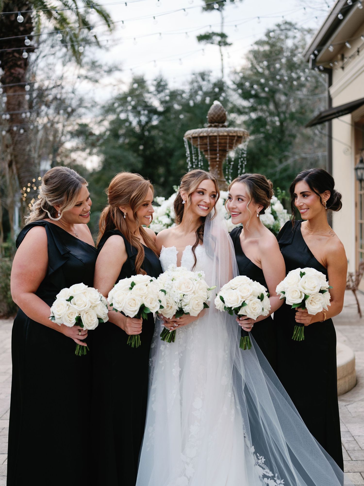 Bridesmaids in elegant black dresses stand together with white bouquets beside a bride in a flowing white gown.