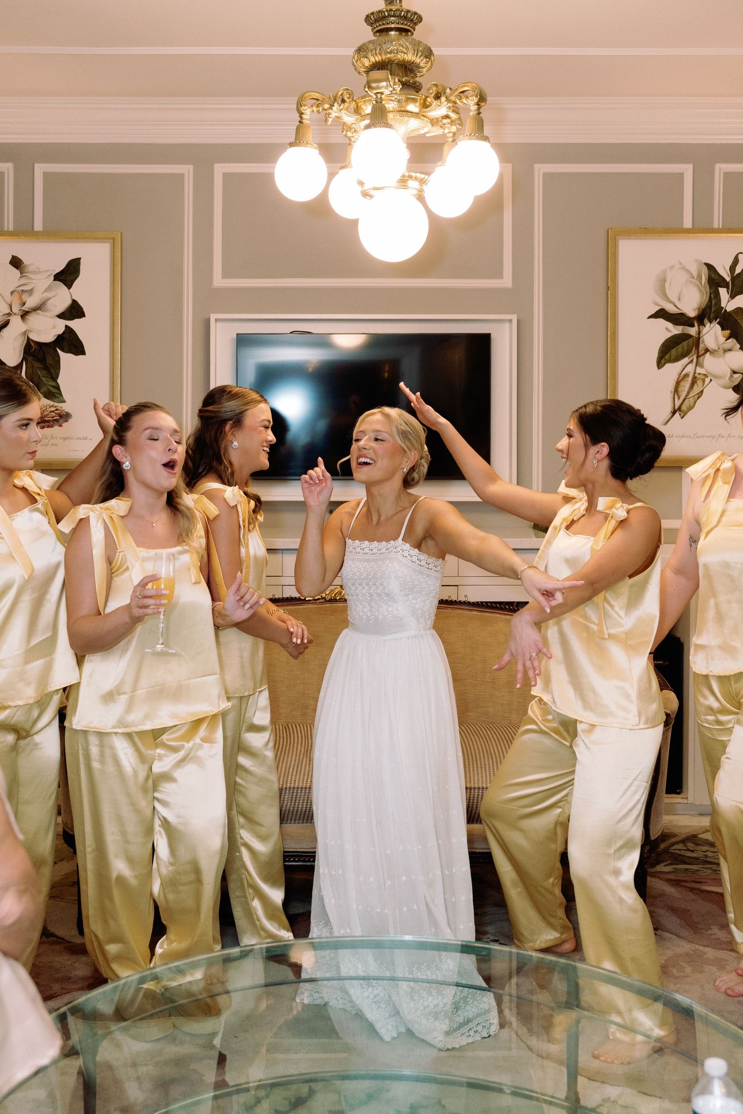 Wedding party in white and champagne dresses dancing joyfully under crystal chandeliers.