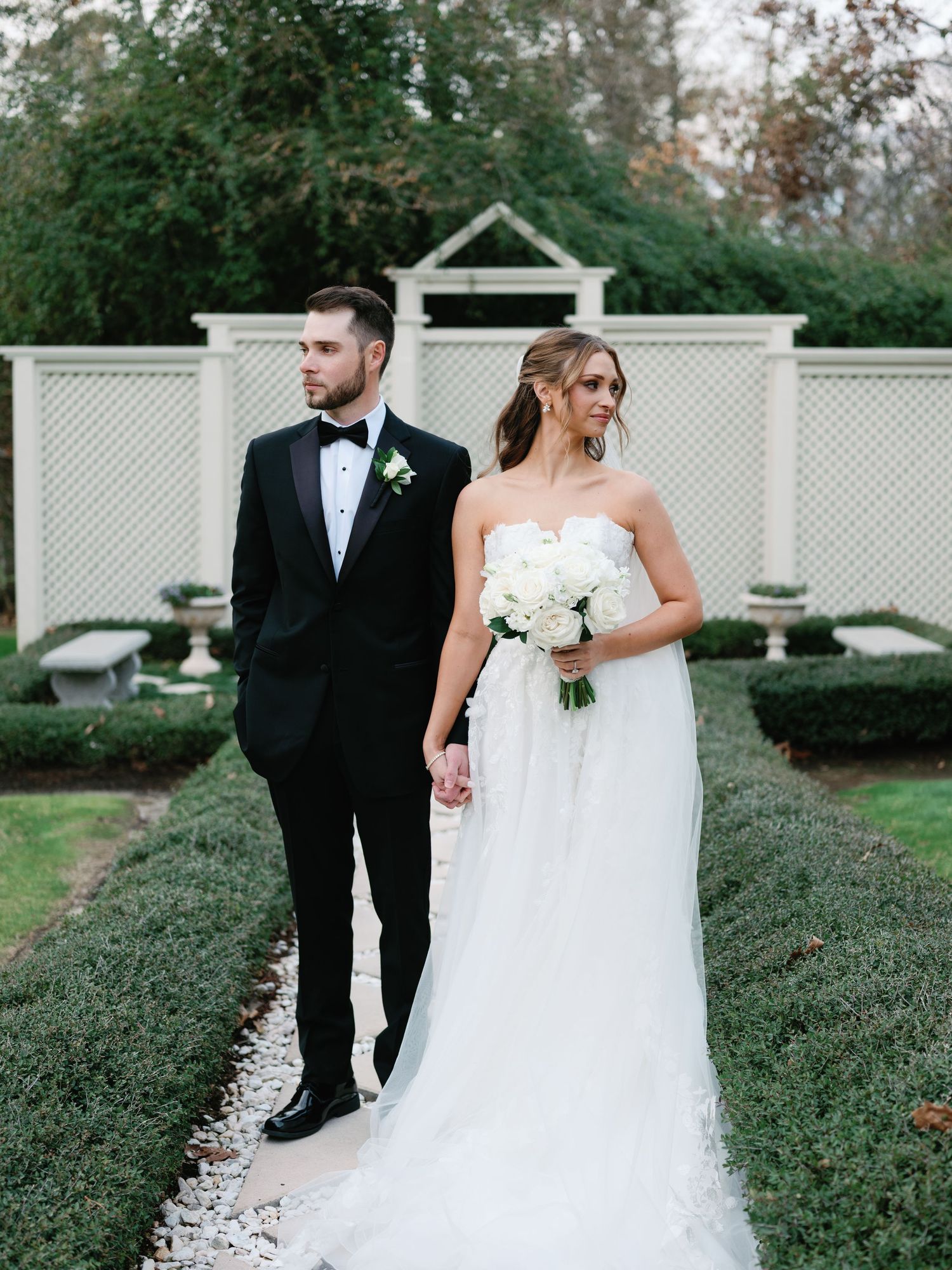 A couple in formal wedding attire poses together in a garden setting with white lattice fencing in the background.