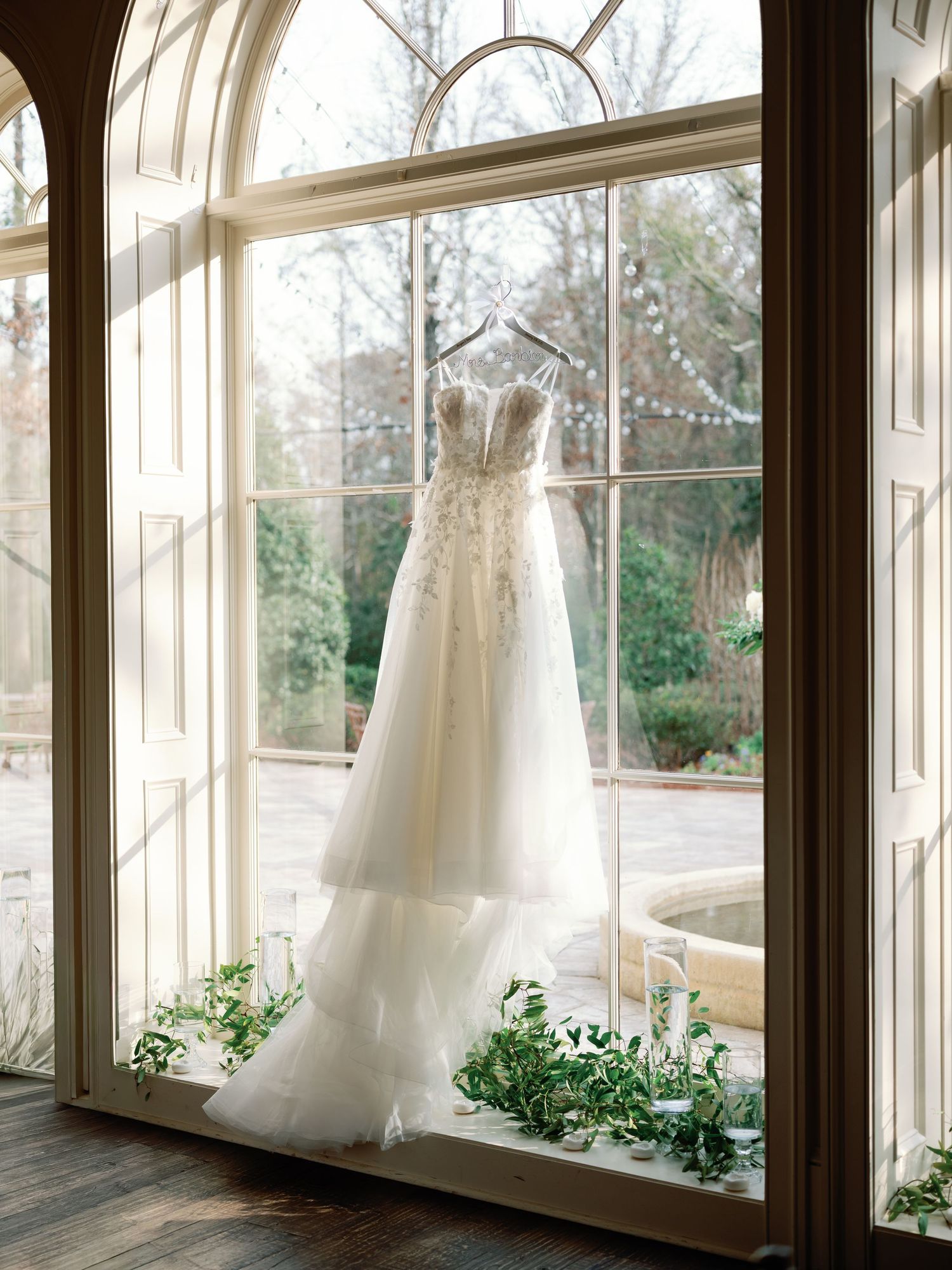 White wedding dresses hanging in sunlit windows with greenery decorations.