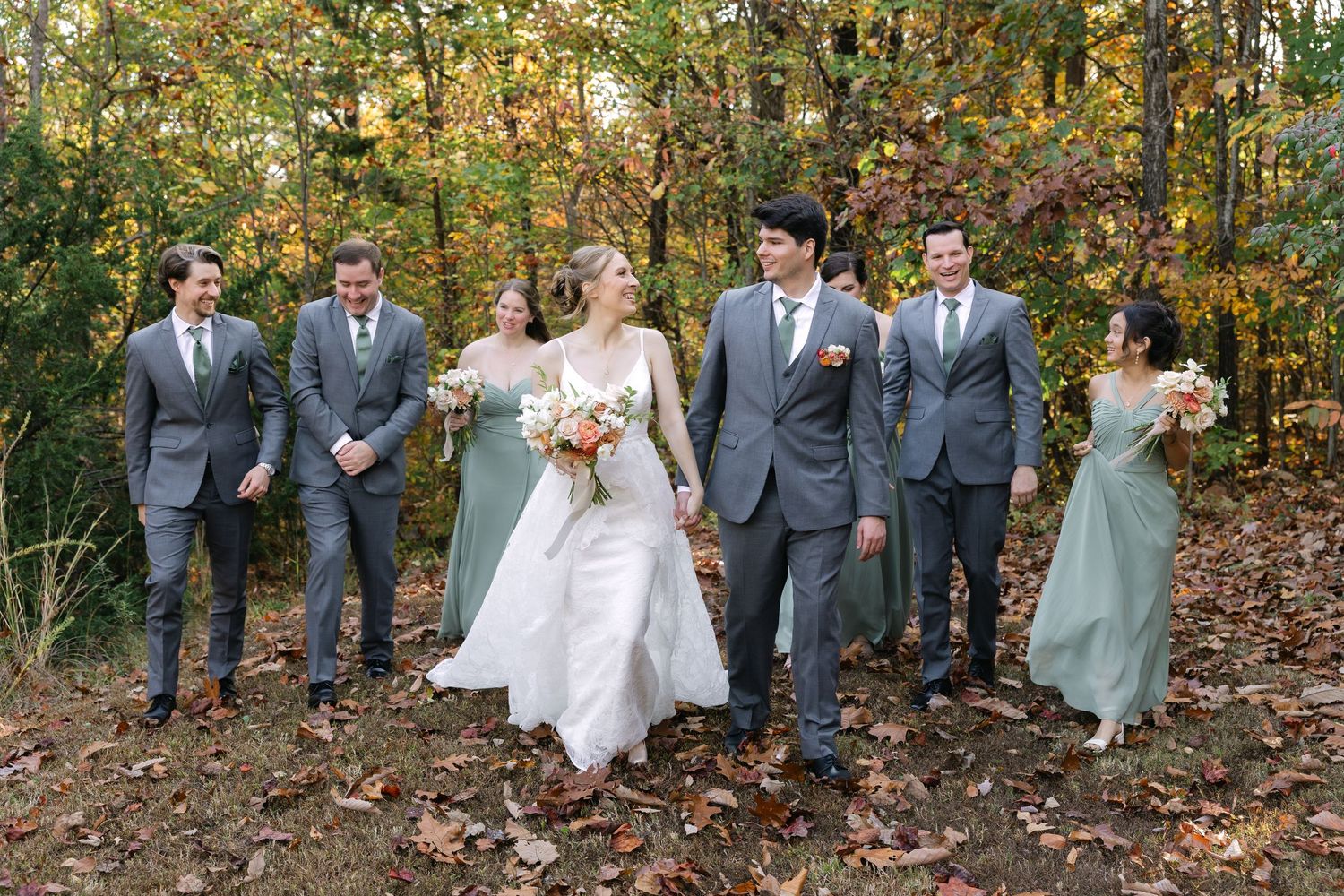 A bride, groom, and their entire wedding party take a walk as they all smile at each other through the woods.