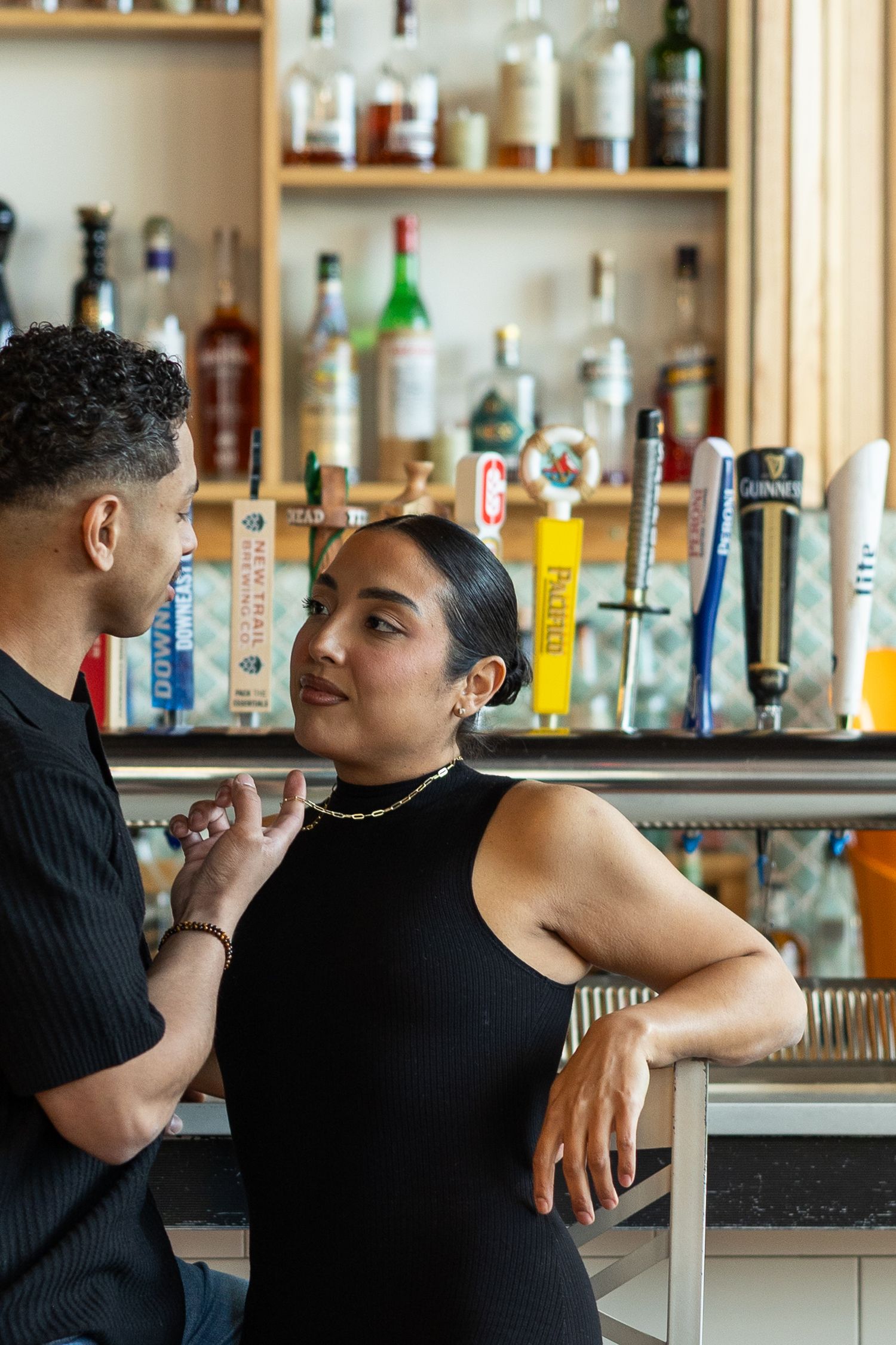 A couple in black outfits chat at a bar with beer taps and liquor bottles on shelves behind them.