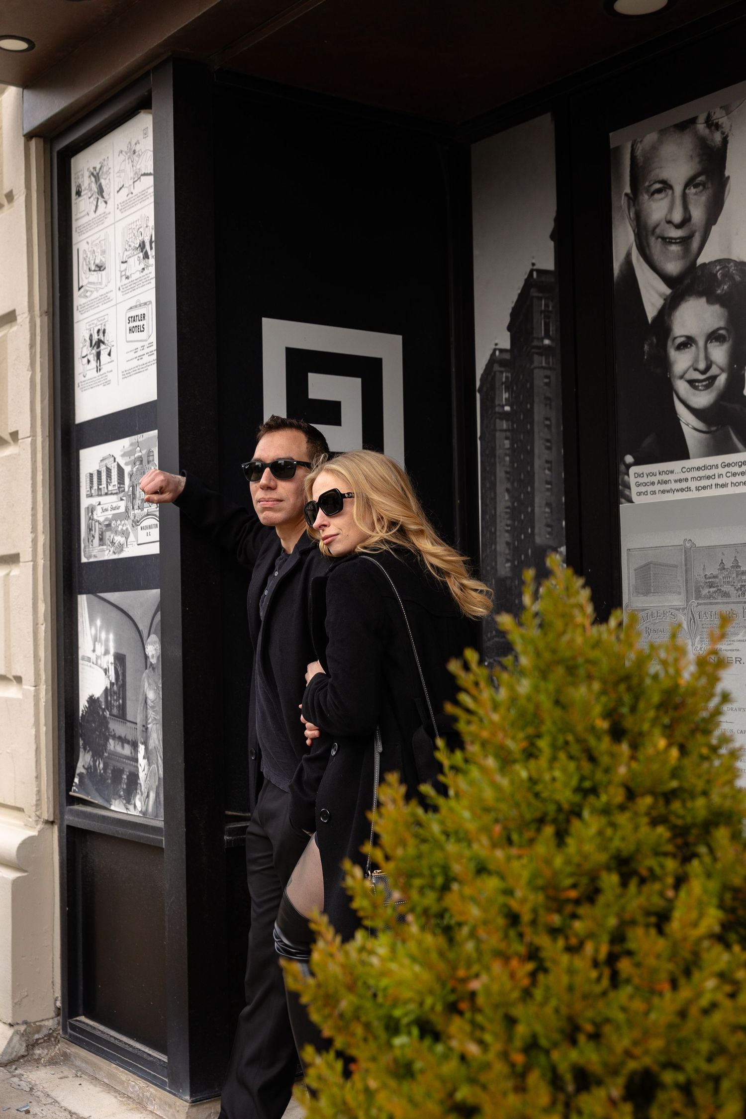 A stylish couple in all-black outfits and sunglasses standing outside a gallery with black-and-white photography displays.