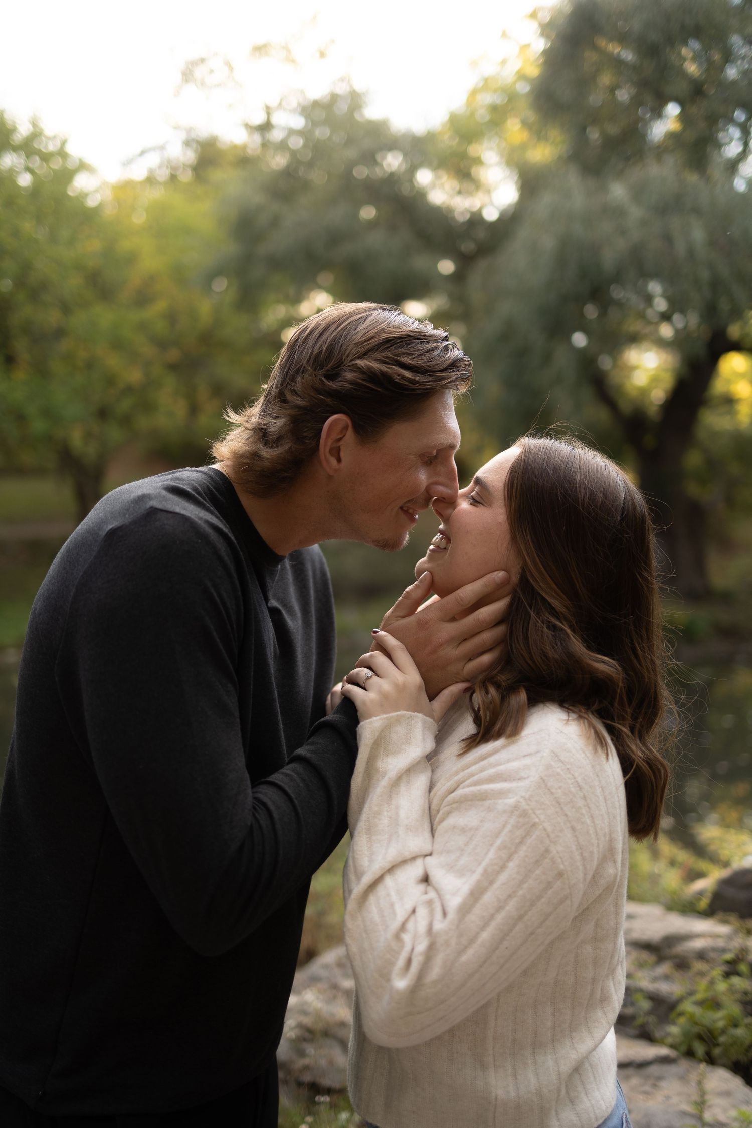 A couple shares a romantic moment in a lush green park, the man tenderly holding the woman's face as they lean in to kiss.