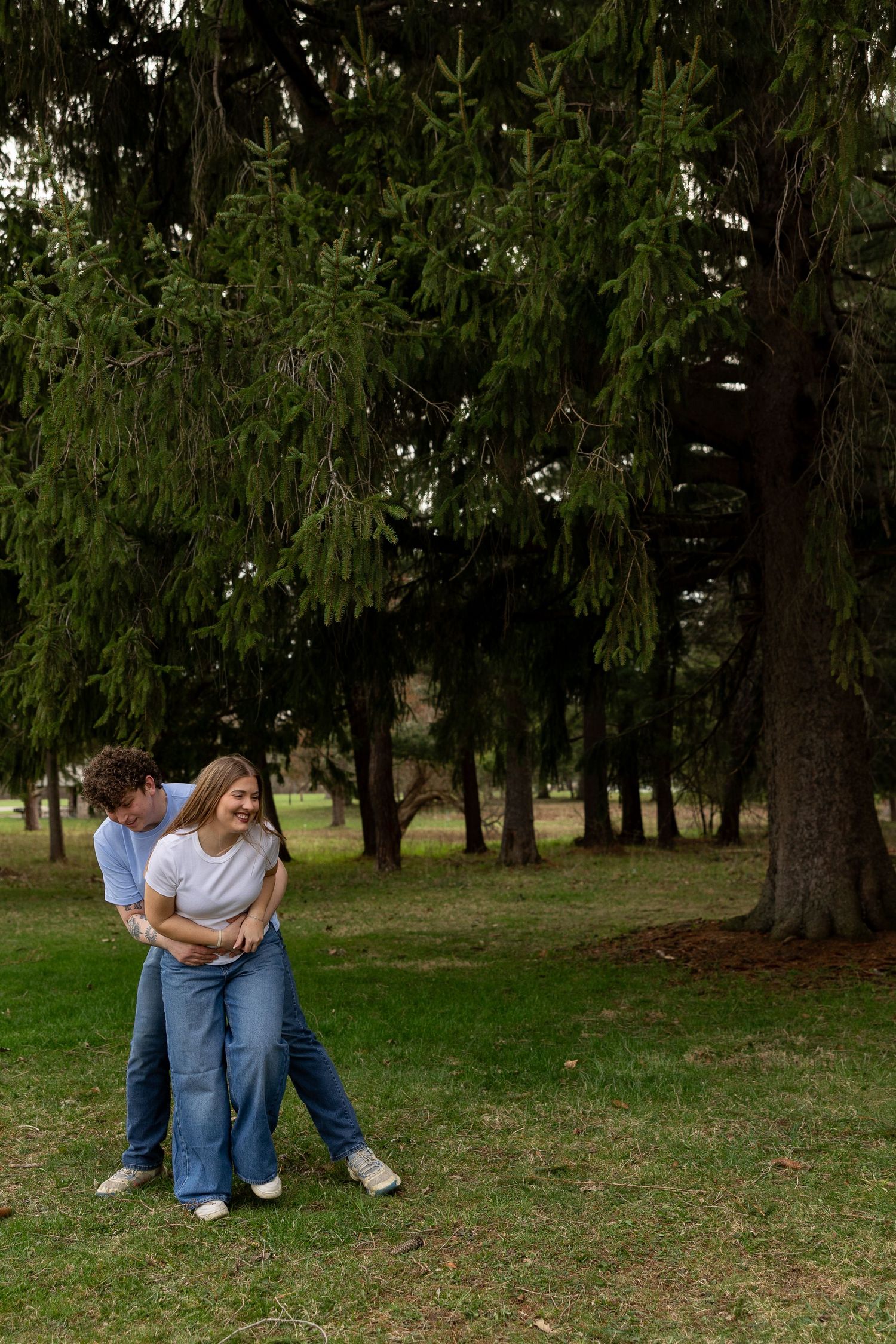 Couple embracing and laughing together in a lush green park surrounded by tall evergreen trees.