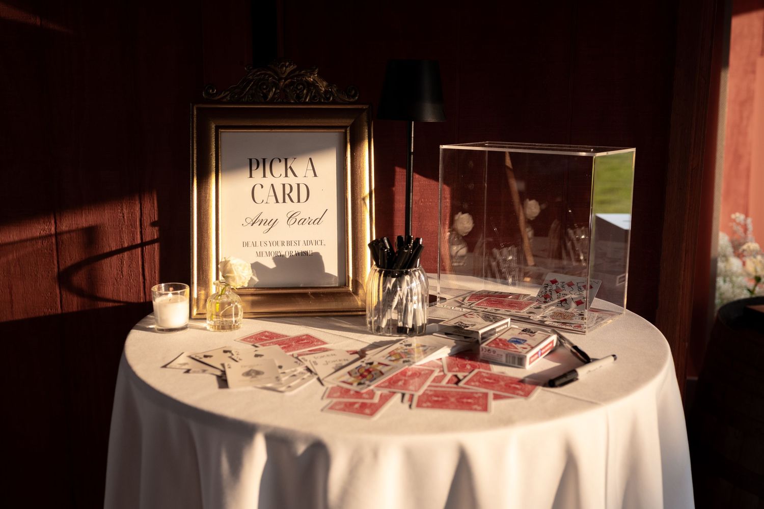 Elegant wedding card table with gold framed Pick A Card sign, playing cards, and white linen in warm lighting.