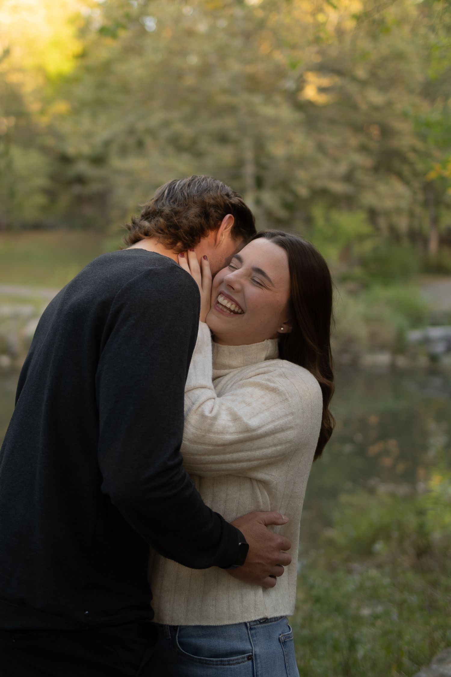 Couple embracing outdoors near a pond, woman laughing joyfully as man kisses her cheek, surrounded by autumn foliage.