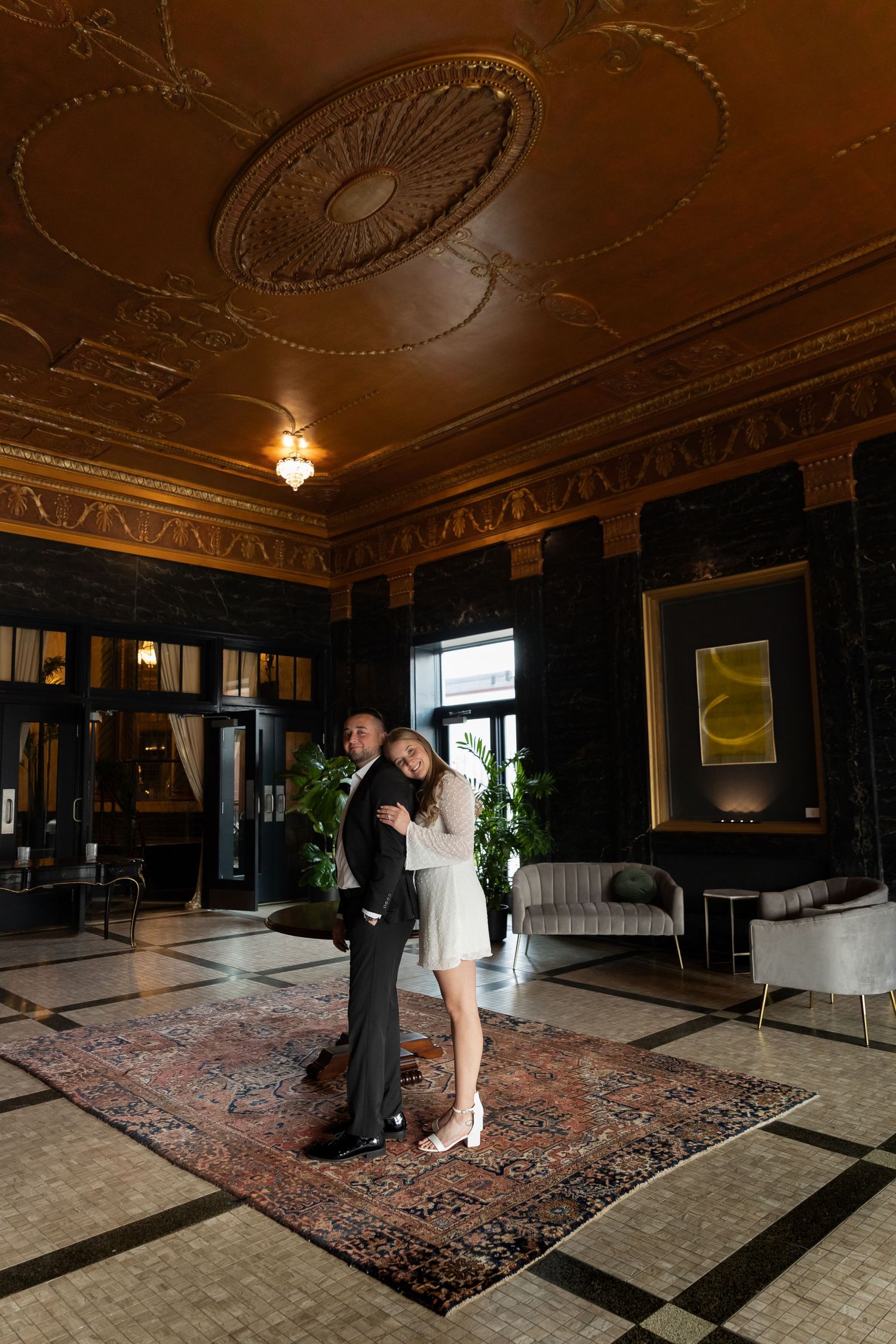 Couple embracing in ornate hotel lobby with gold ceiling, patterned rug, and elegant dark decor.