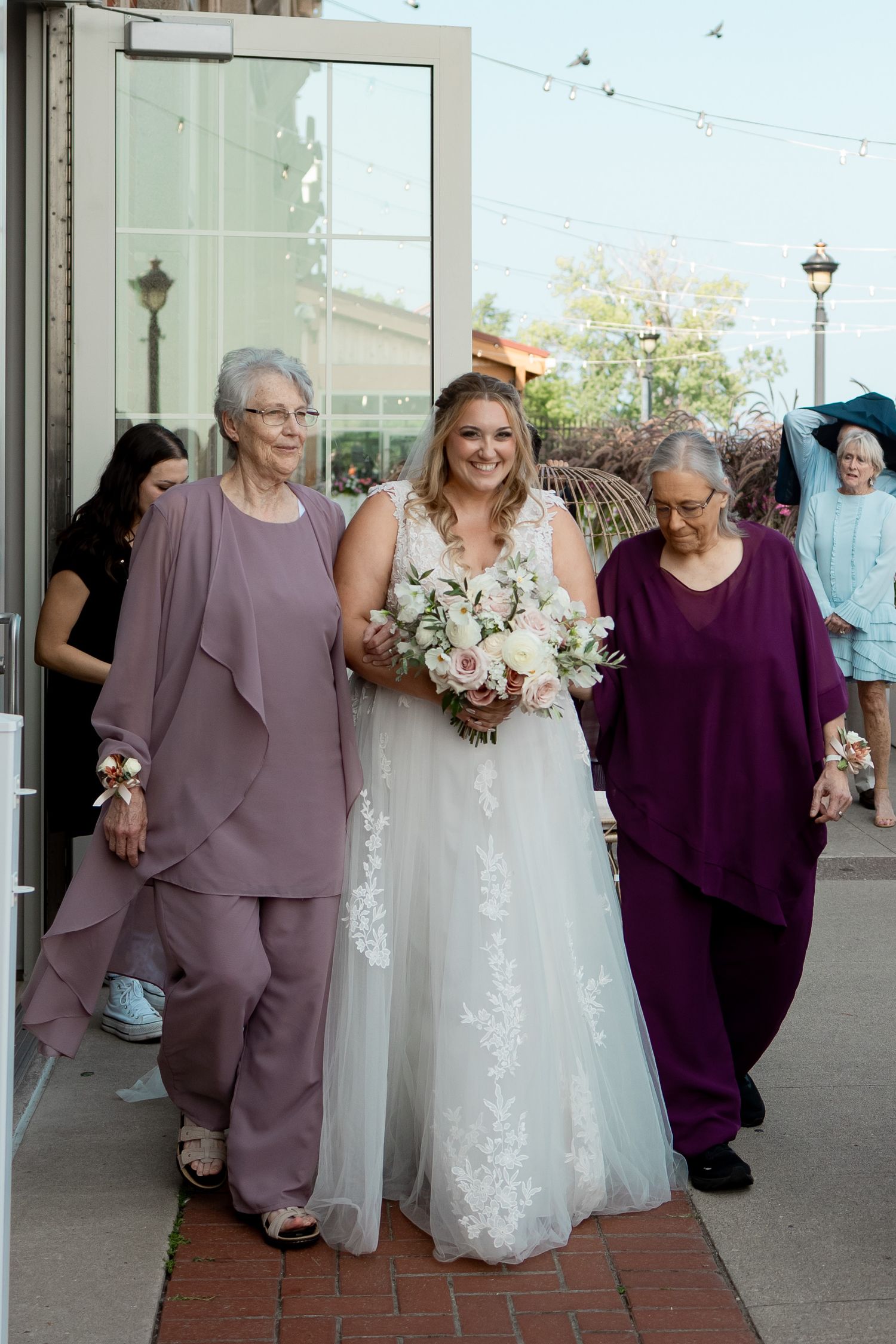 Bride in white lace gown walks with two elderly women in purple outfits, holding a bouquet at an outdoor wedding.