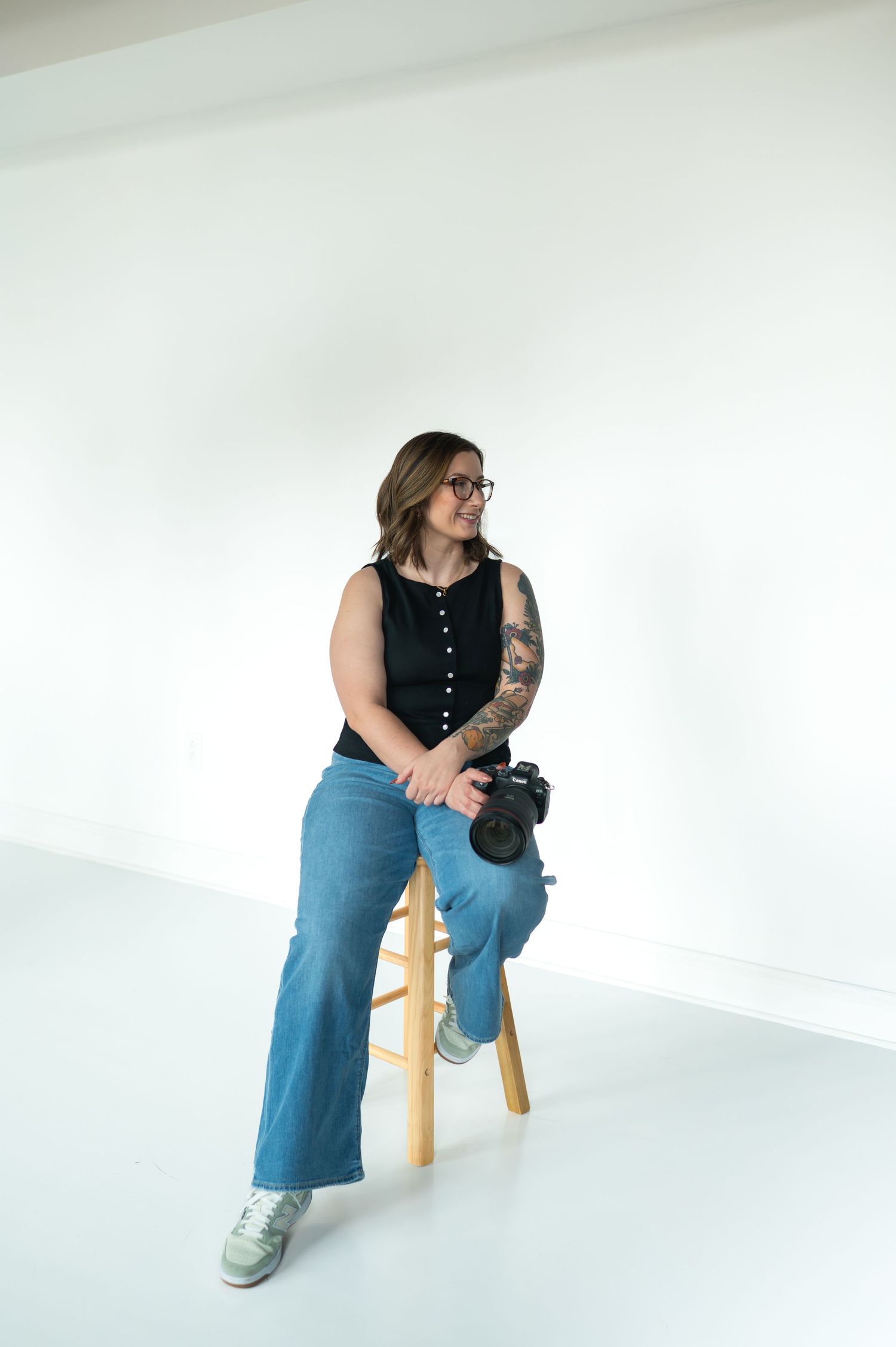 Wedding and Couples Photographer with tattoos sits on stool holding camera in white studio, wearing black top and jeans.