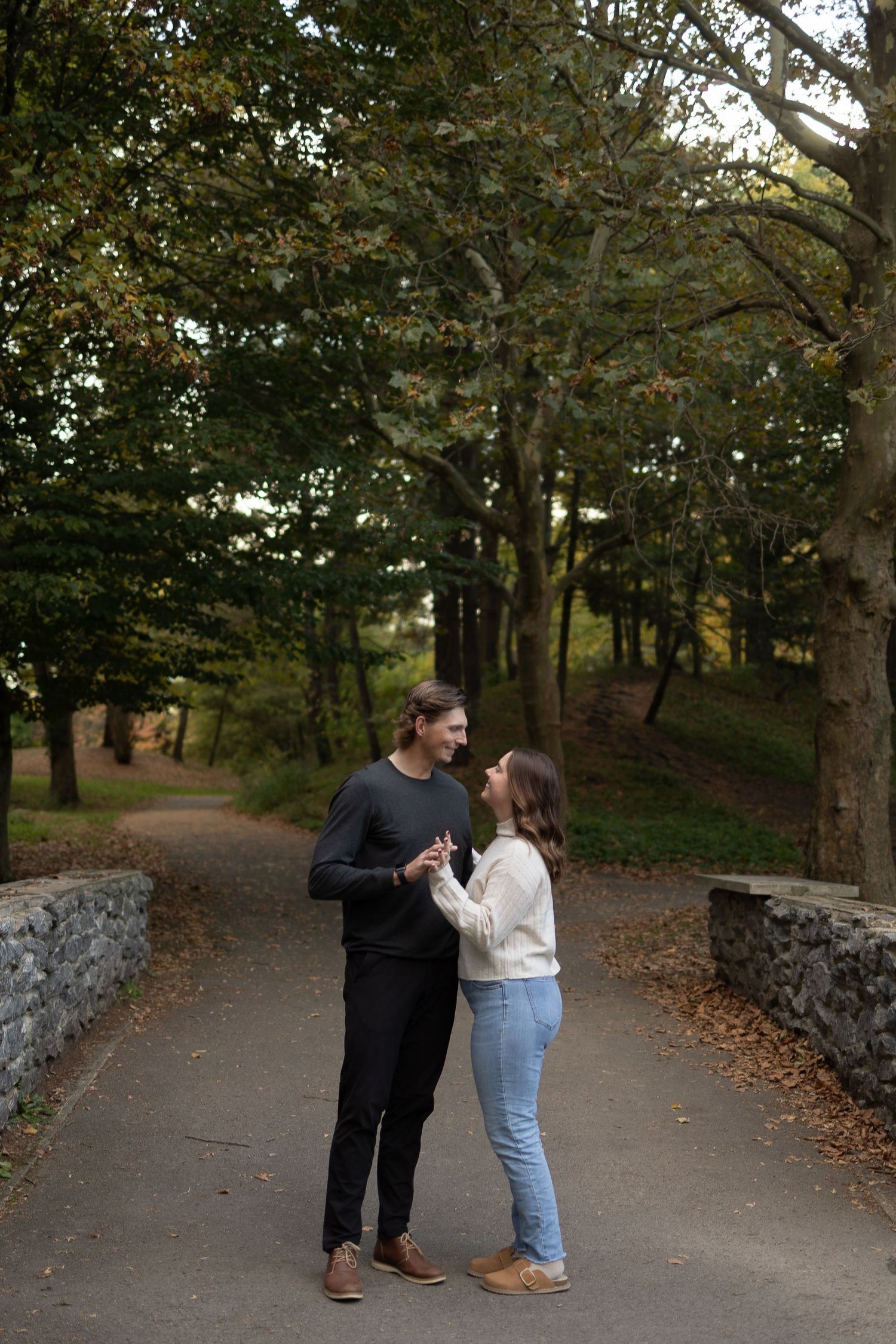 Couple holding hands on a stone bridge path surrounded by lush green trees in an autumn park setting.