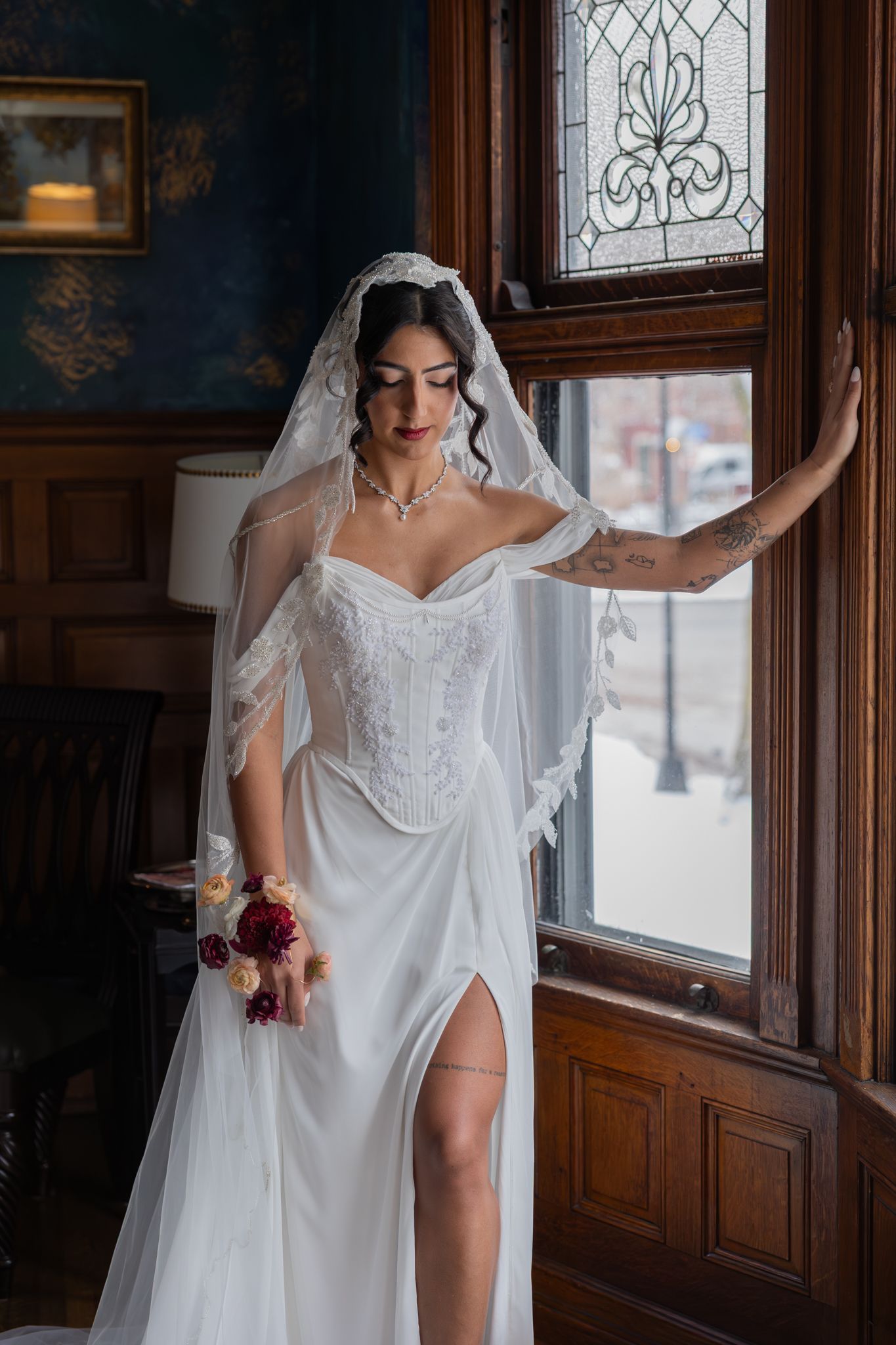 Bride in white corset gown and lace veil stands by ornate wooden window holding burgundy bouquet in elegant Victorian interior.