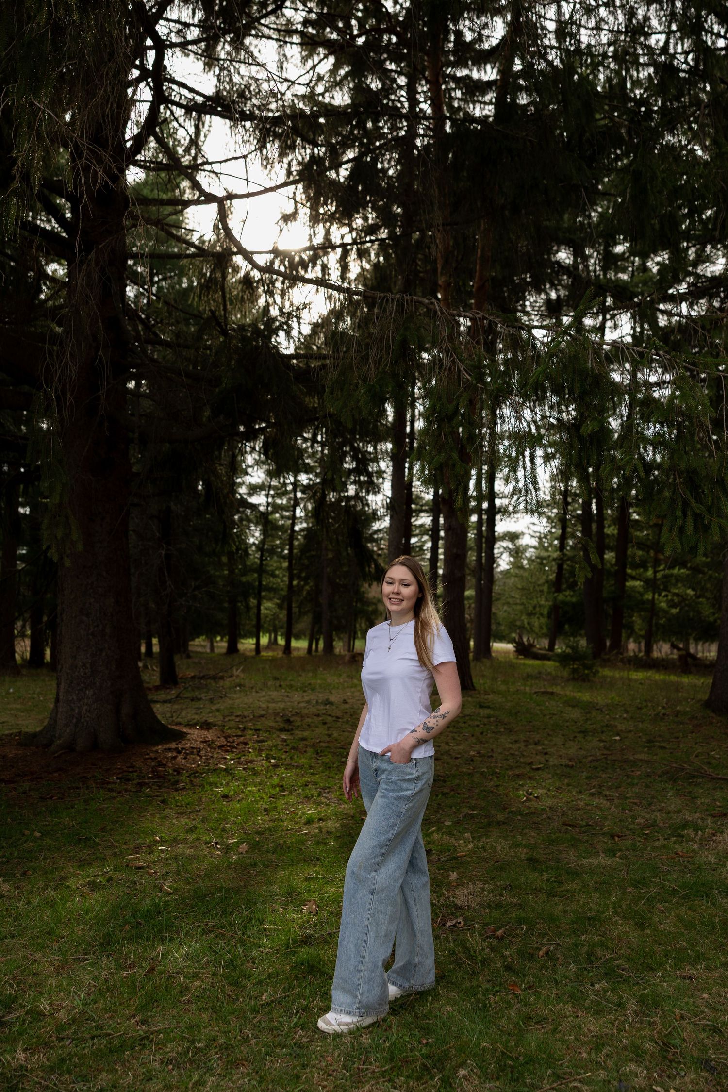 Young woman in white shirt and wide-leg jeans smiling in a lush green forest park setting in Western New York