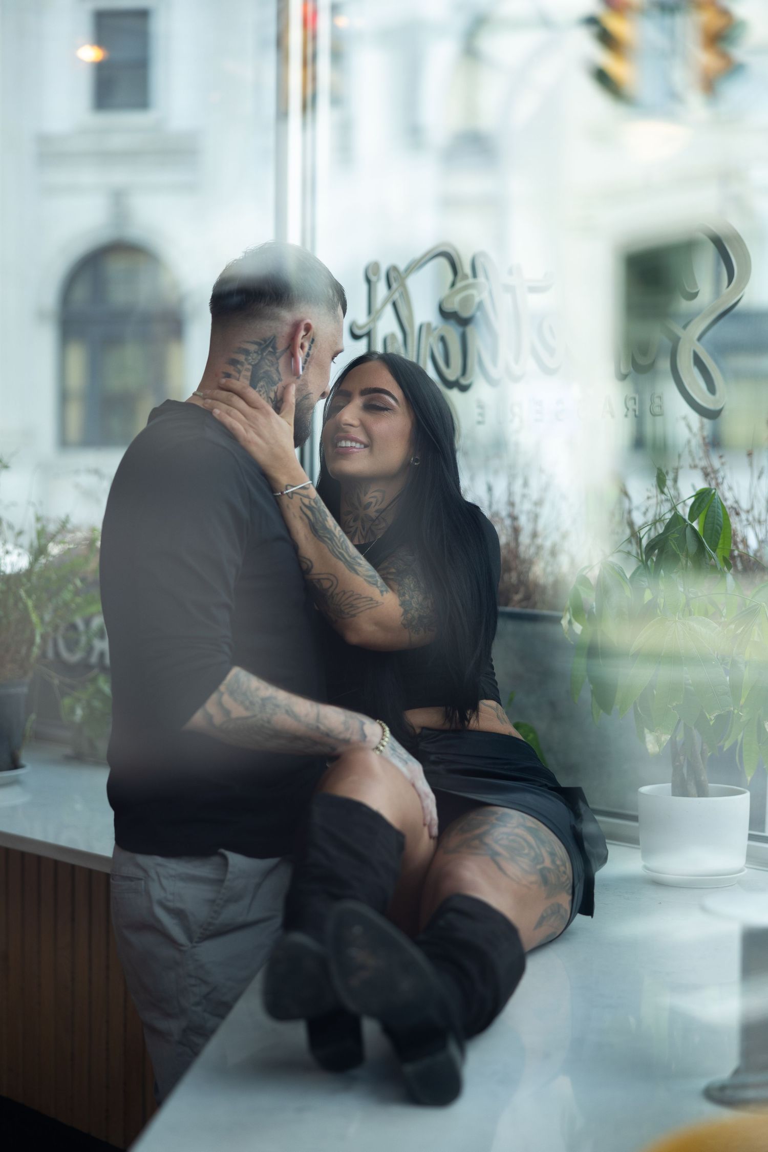 Tattooed couple sharing an intimate moment inside a cafe, woman in black outfit sitting close to man, smiling tenderly.