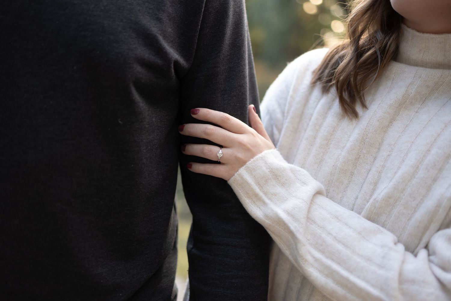A woman wearing an engagement ring holds a man's arm, showcasing a romantic couple moment outdoors.