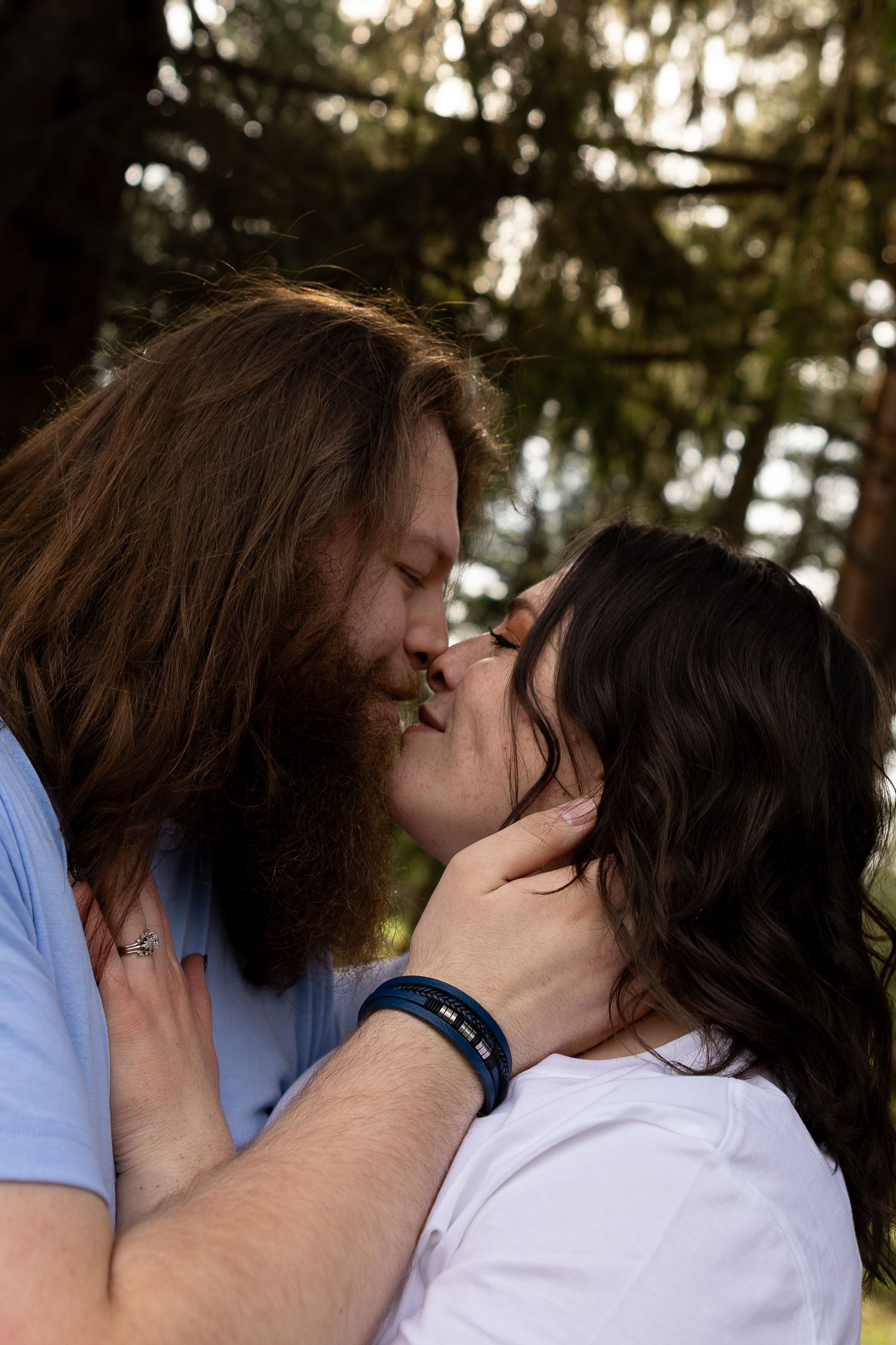 A couple shares a romantic kiss outdoors, surrounded by trees, as the man gently cups the womans face.