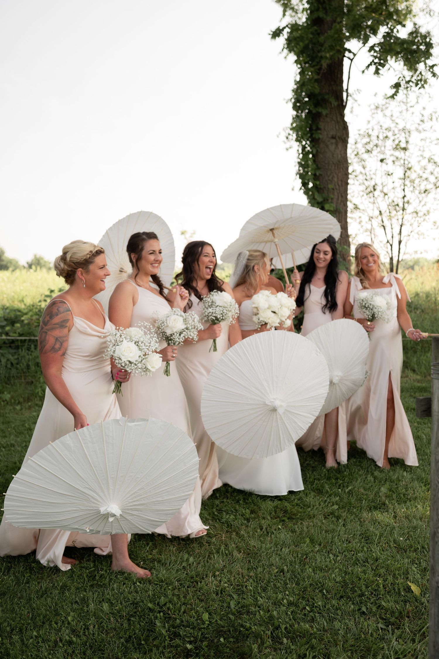 Bridesmaids in blush gowns holding white parasols and floral bouquets stand outdoors on green grass near trees.
