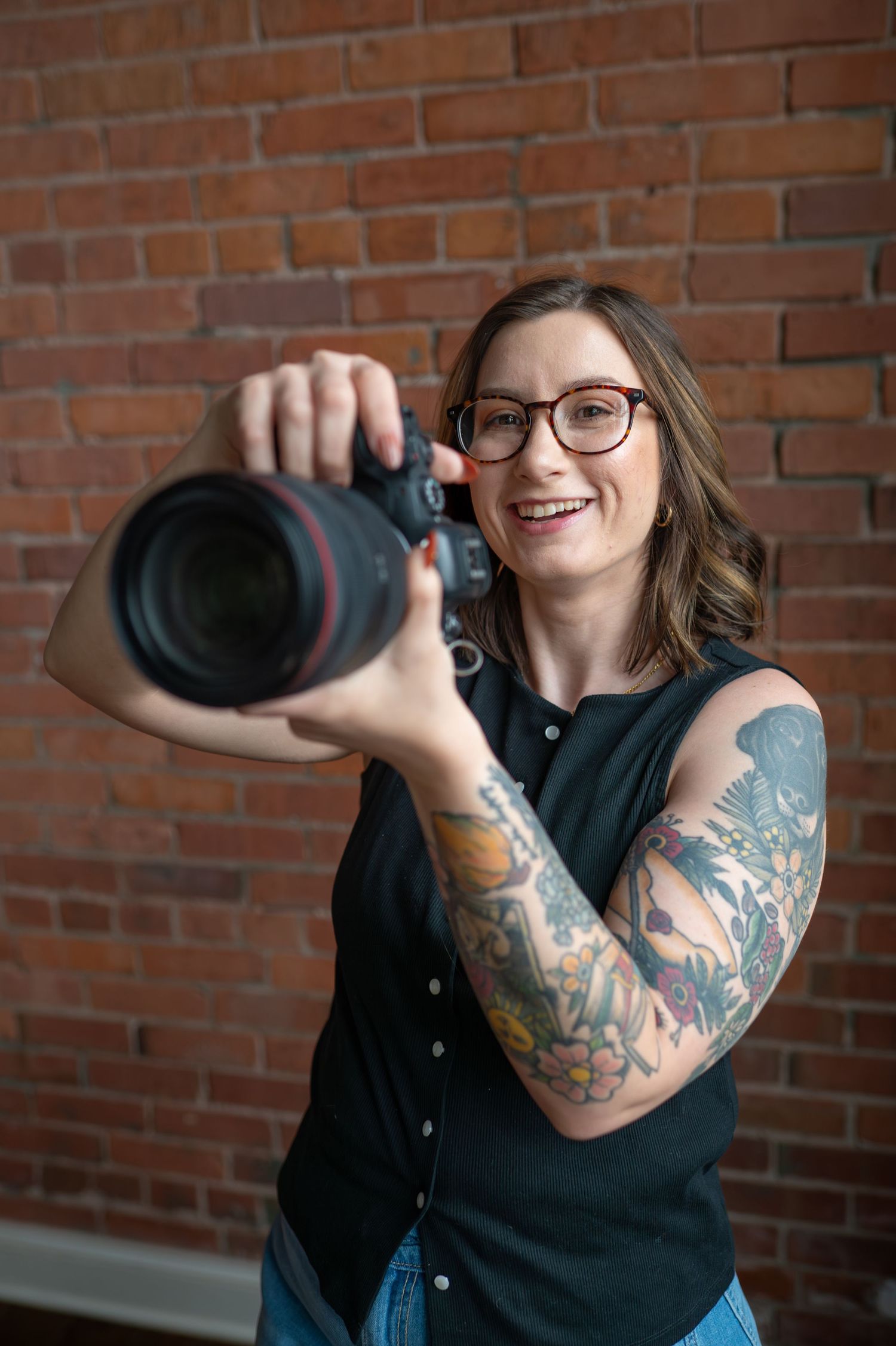 Smiling woman with tattoos and glasses holding a DSLR camera in front of a red brick wall.
