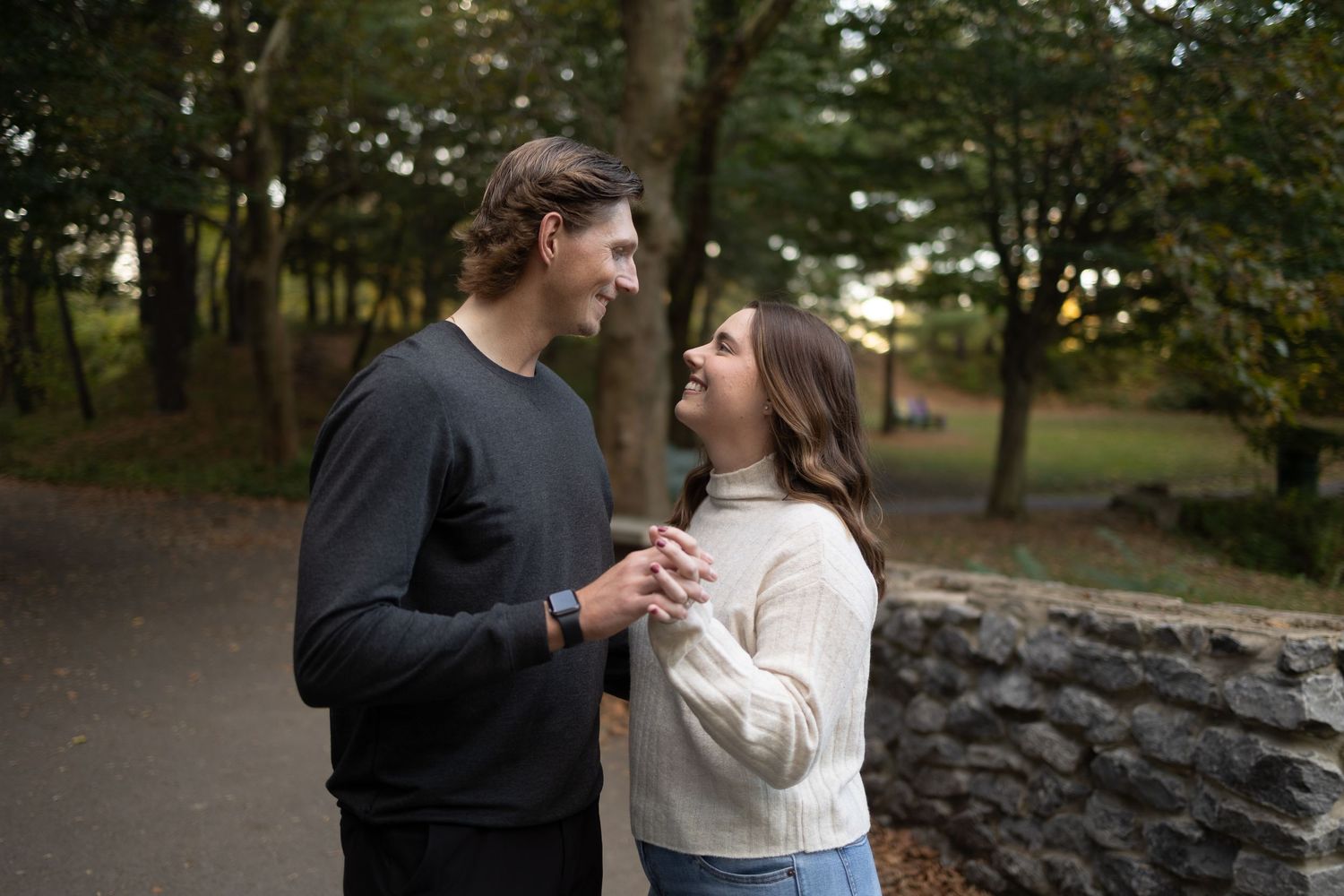 Couple smiling and holding hands at Glen Falls Park surrounded by trees, wearing casual sweaters in autumn.