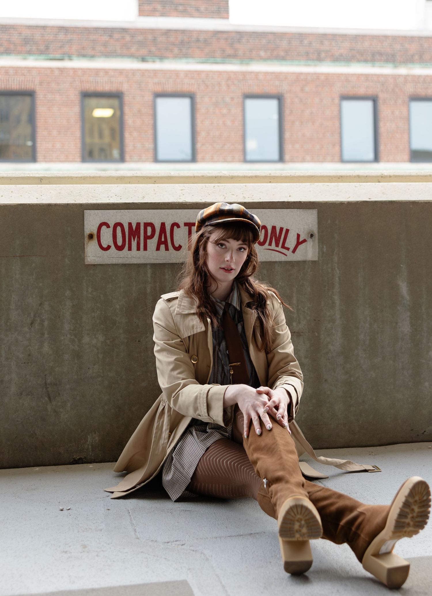 Woman in trench coat, newsboy cap, and knee-high boots sits against a wall near a Compact Cars Only sign.